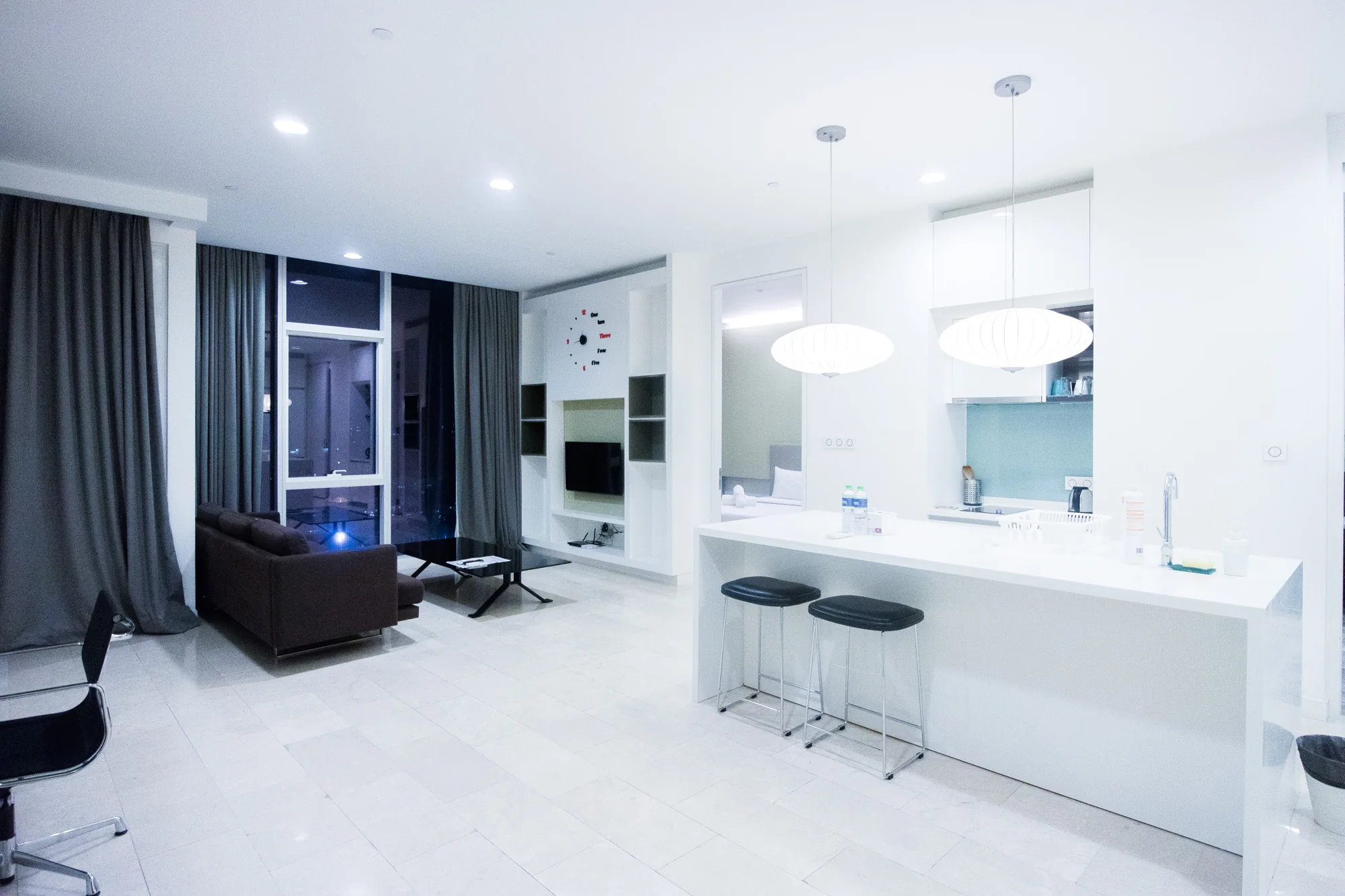 写真はマレーシアのクアラルンプールで撮影された、非常にモダンで明るい室内の様子を捉えています。全体的に白を基調とした内装で、清潔感と広々とした空間が強調されています。床は大理石調の白いタイルで覆われており、光を反射してさらに明るさを増しています。  手前にはキッチンカウンターがあり、2つの黒いスツールが並んでいます。カウンターの上にはボトルや洗剤などが見え、生活感を感じさせます。カウンターの奥にはシンクや調理器具が置かれており、コンパクトながらも機能的なキッチンであることがわかります。  奥にはリビングスペースが広がっており、ダークブラウンのソファと黒いガラステーブルが配置されています。大きな窓からは夜景が望め、グレーのドレープカーテンがエレガントさを添えています。壁には装飾的な時計や棚があり、テレビが設置されています。  全体的にスタイリッシュで洗練された雰囲気であり、高級感のある都会的な暮らしを想像させます。ミニマムなデザインでありながら、必要なものが揃っており、快適な空間であることが伝わってきます。