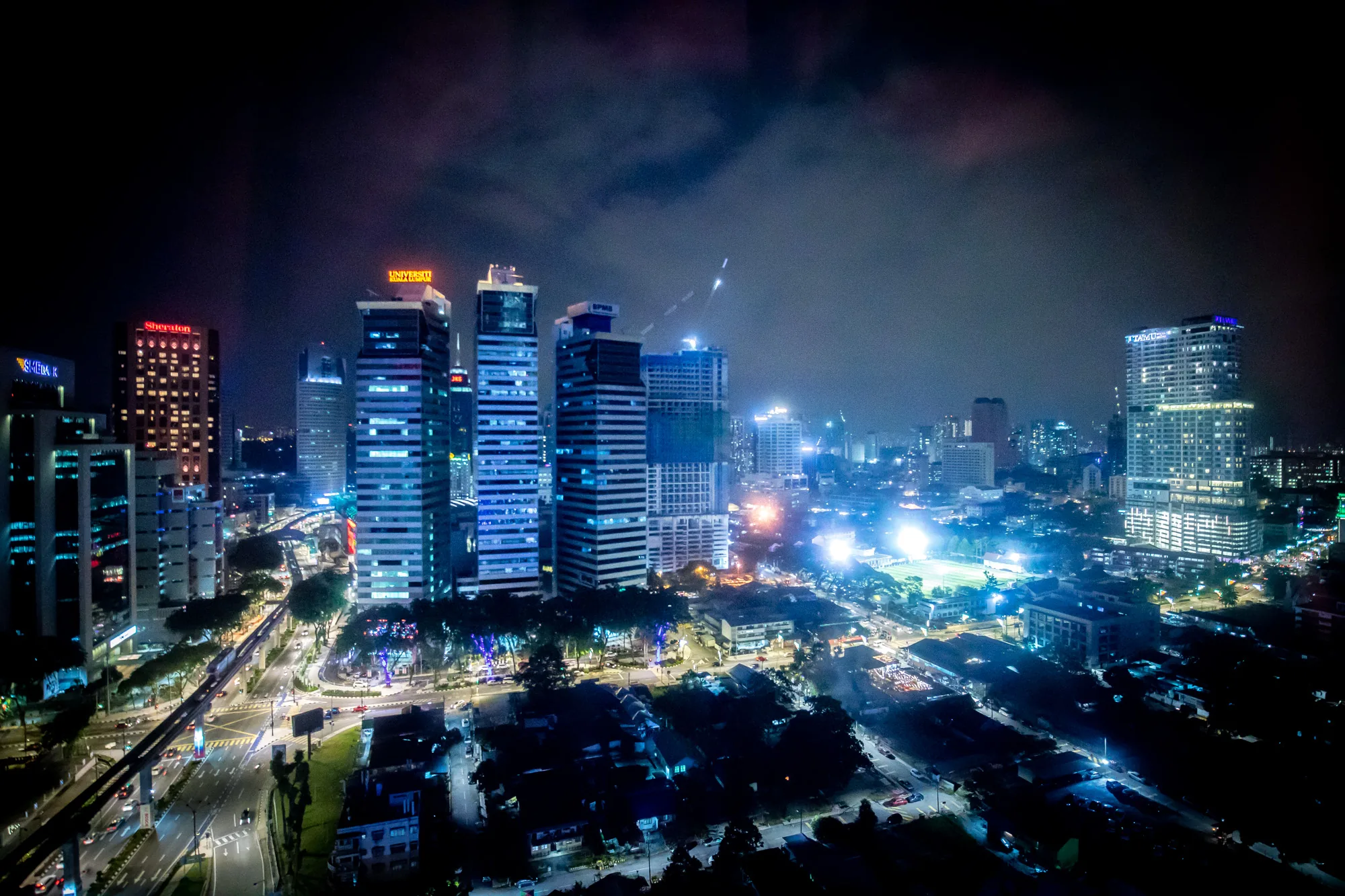 写真はマレーシアのクアラルンプールで撮影された夜景です。高層ビル群がライトアップされ、街全体が明るく照らされています。空には薄く雲がかかっており、幻想的な雰囲気を醸し出しています。手前には住宅街が広がり、遠くに連なるビル群とのコントラストが美しいです。道路には車の光が流れ、街の活気が感じられます。特に目立つのは、いくつかの高層ビルに掲げられた企業ロゴやブランド名（例えば「Sheraton」や「Universiti Kuala Lumpur」）です。これらの光景が、クアラルンプールの都会的な魅力を伝えています。