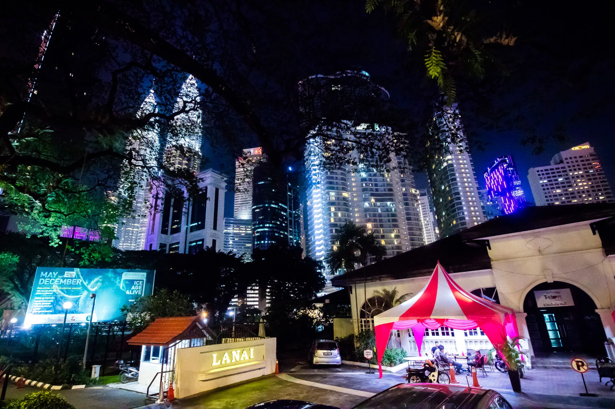 この写真は、マレーシアのクアラルンプールで撮影された夜景です。前景には「LANAI」と書かれた看板のある建物があり、入り口には赤と白のストライプのテントが設置されています。テントの下には人が何人か座っています。背景には高層ビル群がそびえ立ち、様々な色でライトアップされています。特に、ペトロナスツインタワーがひときわ目立っています。写真全体は暗い夜空と建物の光によって照らされており、都会的で賑やかな雰囲気を醸し出しています。道路沿いには看板があり、「MAY-DECEMBER」「ICE AGE ALIVE」などの文字が見えます。夜のクアラルンプールの活気が感じられる一枚です。