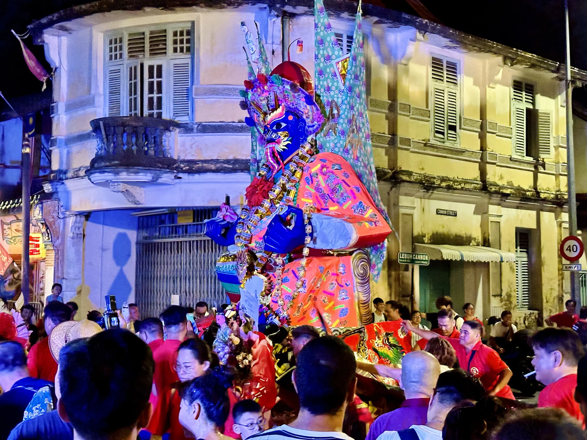 これはマレーシアのペナンで撮影されたと思われる夜間の写真です。画面の中央には、鮮やかな青い顔と派手な装飾を施された神輿のようなものが置かれ、その周囲には多くの人々が集まっています。人々は神輿を見上げたり、写真を撮ったりしている様子です。参加者はほとんどが赤色のシャツを着用しているようです。背景には、コロニアル様式の建物が並び、ペナンの歴史的な街並みを彷彿とさせます。建物はライトアップされ、独特の雰囲気を醸し出しています。街灯や店の明かりが暗闇を照らし、祭りの活気と興奮を伝えています。全体的に、エネルギッシュで文化的な雰囲気が感じられる写真です。