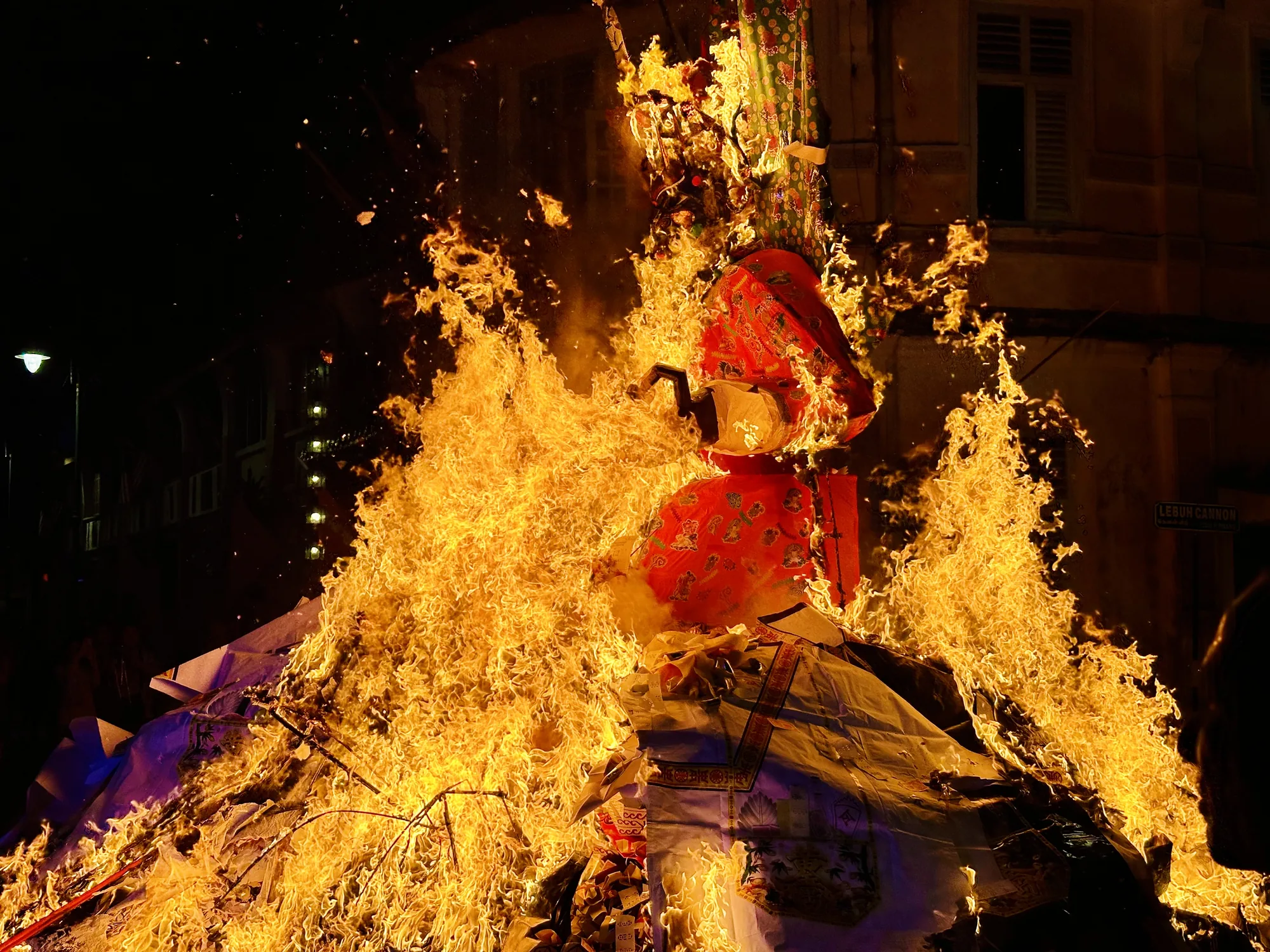 マレーシアのペナンで撮影された写真です。暗闇の中で、巨大な炎が燃え盛っている様子が捉えられています。炎の中心には、布で覆われた人形のようなものが立っており、その周囲を炎が激しく包み込んでいます。人形の上部には、緑色の花柄の布が、下部には赤い布が掛けられています。炎の勢いが非常に強く、火の粉が舞い上がり、熱気が伝わってくるようです。背景には、古い建物の一部が見え、窓や壁の質感が確認できます。看板には「LEBUN CANNON」と書かれています。全体的に、神秘的で力強い雰囲気を感じさせる一枚です。お祭りか何かの儀式の様子を捉えたものと考えられます。