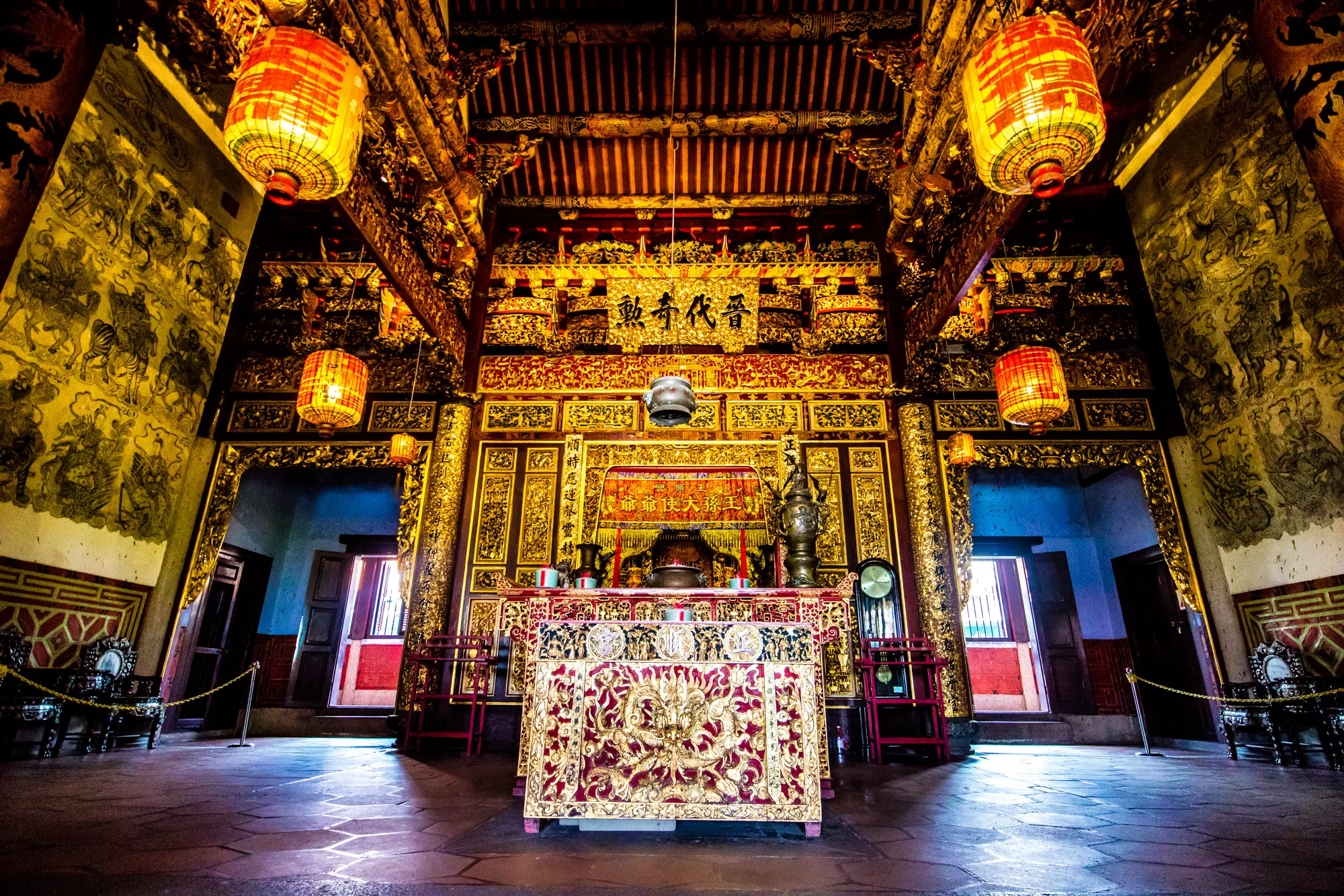 写真はマレーシアのペナンにある寺院の内部を写したものです。画面中央には豪華な装飾が施された祭壇が配置されています。祭壇の前には、金色の装飾が施された赤いテーブルが置かれ、その上には香炉や供え物が置かれています。祭壇の背後には、金色の彫刻が施された壁があり、漢字が書かれた額が掲げられています。  天井は木製の梁が組まれ、同じく金色の装飾が施されています。天井からは、赤と黄色の提灯が吊り下げられています。壁には、人物や動物が描かれた壁画が見られます。部屋の左右には、金色の装飾が施された柱があり、豪華な雰囲気を醸し出しています。  部屋全体は、荘厳で神聖な雰囲気に包まれています。祭壇の細部に至るまで丁寧に施された装飾や、壁に描かれた壁画から、この寺院が長い歴史と伝統を持っていることが伺えます。