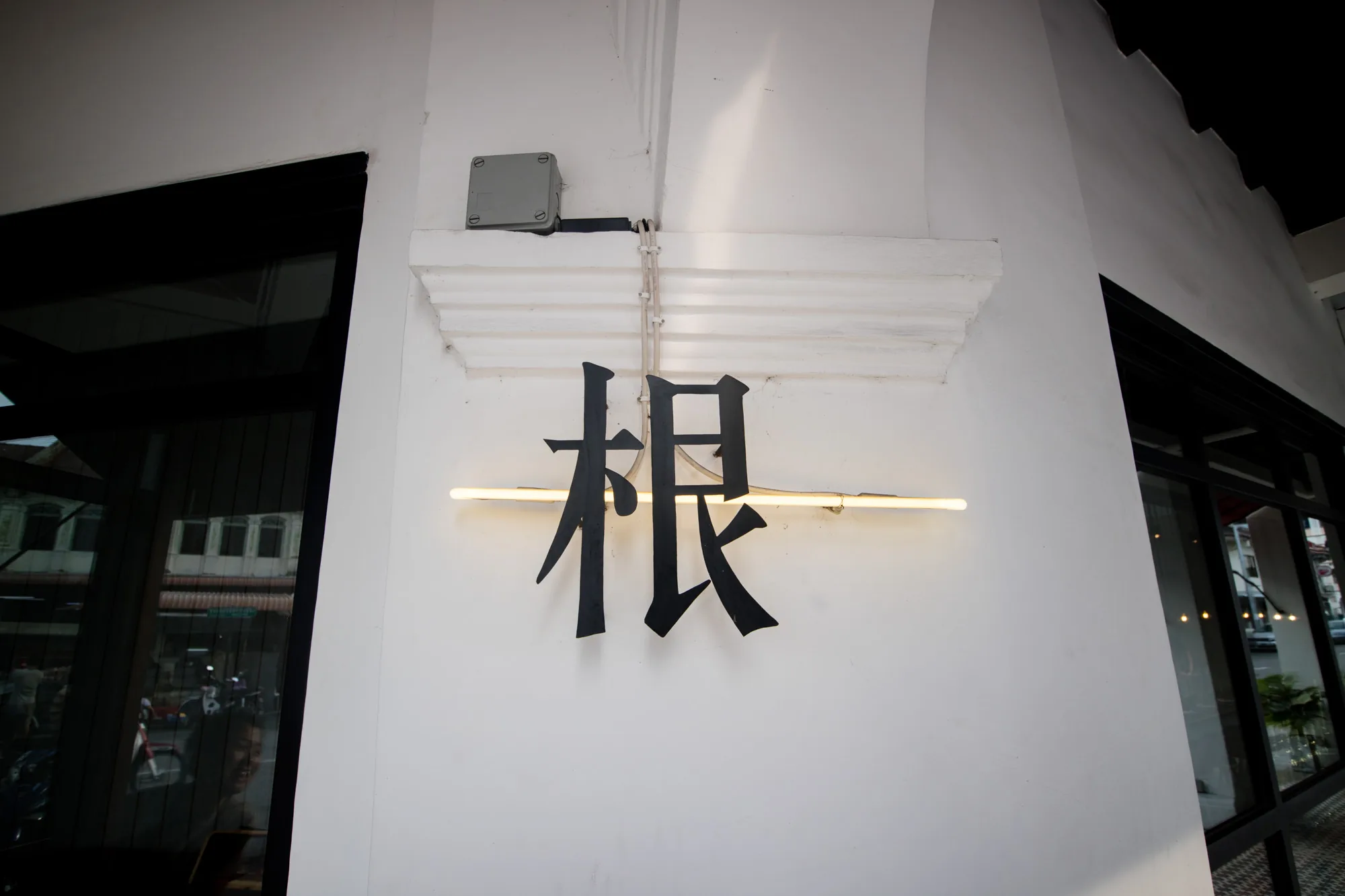 写真はマレーシアのペナンで撮影されたと思われるもので、白い壁に黒い漢字一文字の看板「根」が取り付けられている様子を捉えています。「根」の字は、背面に設置された蛍光灯によって照らされており、モダンで洗練された印象を与えます。壁の上部には、建物の装飾的な白いモールディングが施されており、電気配線が露出しています。左側には黒い枠の窓ガラスが写り込んでおり、外の街並みや人物がぼんやりと映っています。全体的に、シンプルながらも印象的な看板のデザインが際立つ写真です。