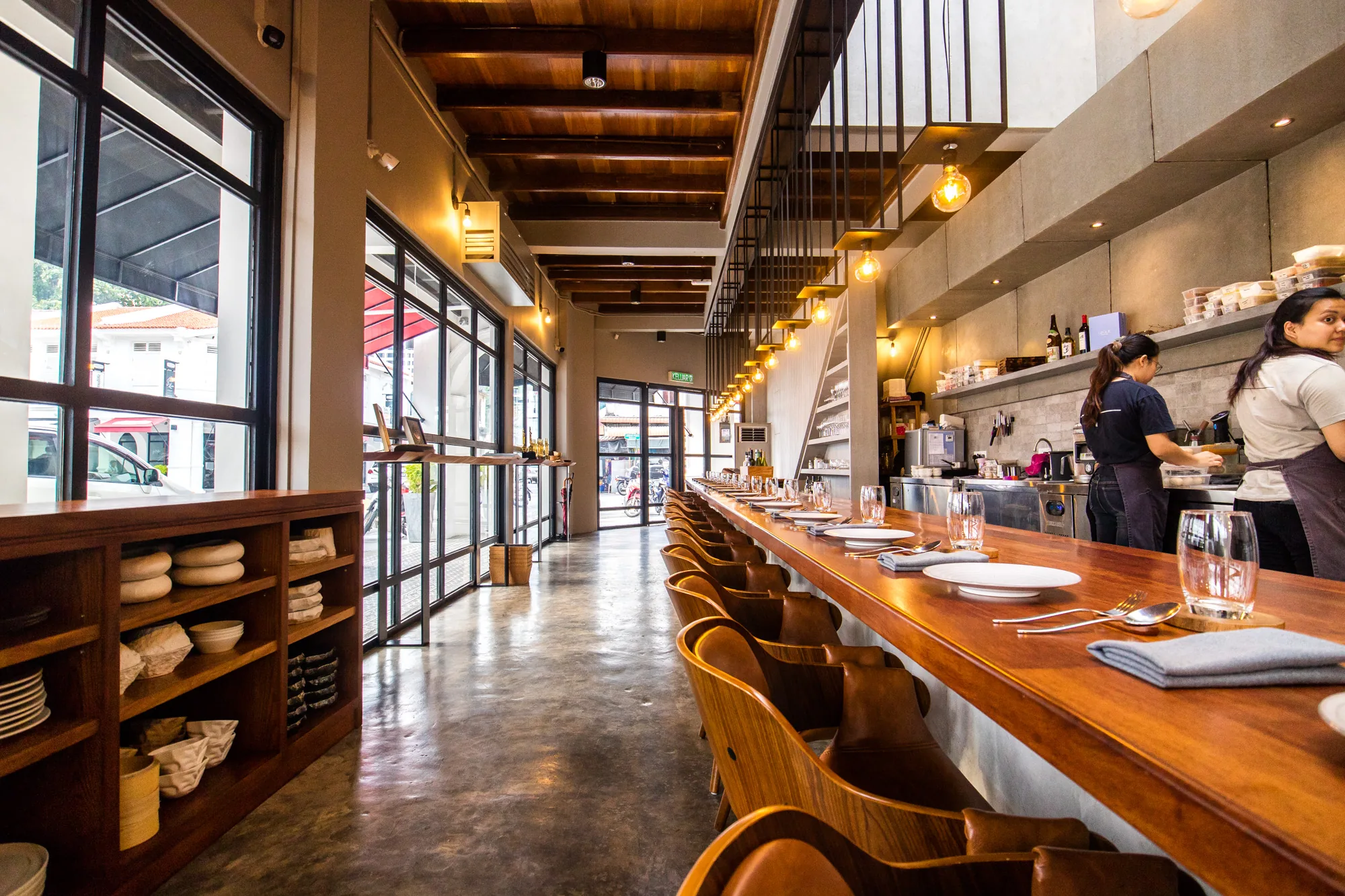 写真は、マレーシアのペナンにあるモダンなレストランのインテリアを写しています。視線は奥へと伸びるように、長いカウンターテーブルと、その前に並んだ曲線的な背もたれが特徴的な木製の椅子に焦点が当てられています。テーブルには、白い皿、グラス、カトラリー、そしてグレーのナプキンがきちんと並べられ、上品な雰囲気を醸し出しています。  写真の右側にはオープンキッチンがあり、エプロンをつけた女性たちが調理作業をしています。背景には、調味料や調理器具などが整理整頓された棚が見えます。天井は木製の梁で装飾され、その下には裸電球が吊り下げられ、温かい光を放っています。  左側には大きな窓があり、外の風景を垣間見ることができます。窓枠は黒で、モダンな印象を与えています。手前には木製の棚があり、様々な食器がディスプレイされています。  全体的に、洗練された雰囲気と温かみのある照明が組み合わさった、居心地の良い空間であることが伝わってきます。レストランは清潔感があり、細部にまでこだわりが感じられます。