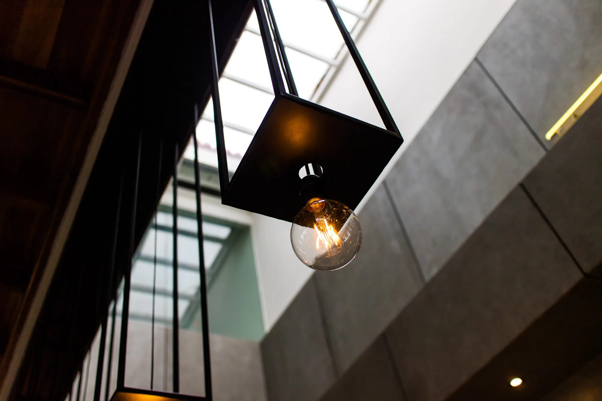 写真は、マレーシアのペナンで撮影された、モダンなインテリアの一部を捉えたものです。主な被写体は、吊り下げられた照明器具で、正方形の黒いフレームの中に、丸い電球が収められています。電球は琥珀色に光っており、内部のフィラメントがはっきりと見えます。フレームは細い黒い線で構成されており、天井に向かって伸びています。背景には、幾何学的な形状の窓や、コンクリートのような質感の壁が見えます。写真全体の色調は、暖色の電球の光と、冷たいコンクリートの灰色が組み合わさって、落ち着いた雰囲気を醸し出しています。照明器具のミニマルなデザインと、背景のモダンな建築様式が調和しており、スタイリッシュな空間を印象づけています。