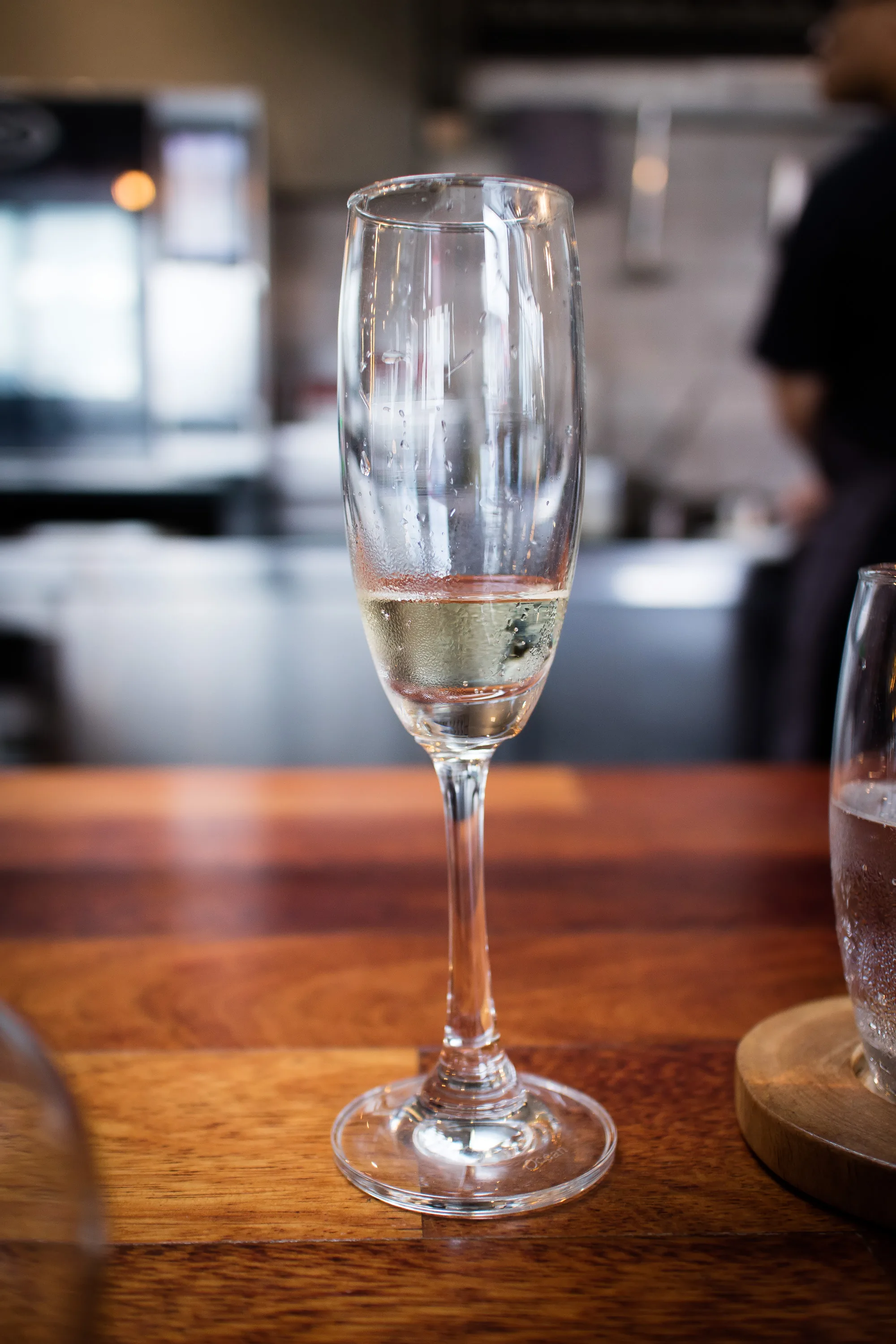 写真には、木製のテーブルの上に置かれたシャンパングラスが写っています。グラスの中にはわずかにシャンパンが残っており、細かい泡が立っています。グラスの内側には水滴が付着しており、冷えていたことが伺えます。背景には、ぼやけていますが、レストランの厨房のような場所が見えます。調理をしていると思われる人物の姿も確認できます。テーブルの木目は美しく、光沢があります。全体的に、食事後のリラックスした雰囲気を感じさせる写真です。