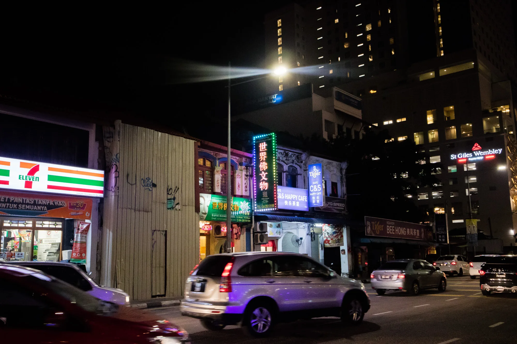 この写真はマレーシアのペナンで撮影された夜の街並みを捉えています。画面左側には、おなじみの7-Elevenの看板が鮮やかに光っています。その隣には、波板のような壁が立っており、落書きが施されています。その奥には、カラフルなネオンサインが輝く商店が軒を連ねています。「世界佛文物」と書かれた看板は特に目を引きます。その他、「Tiger」ビールやカフェの看板も見えます。  道路には車が何台か走っており、その光跡が動きを感じさせます。奥には、St Giles Wembleyというホテルが見えます。街灯が強く光を放ち、夜の街を照らしています。全体的に、ペナンの活気ある夜の雰囲気を伝える写真です。新旧の建物が混在している様子や、様々な言語の看板が、異国情緒を醸し出しています。