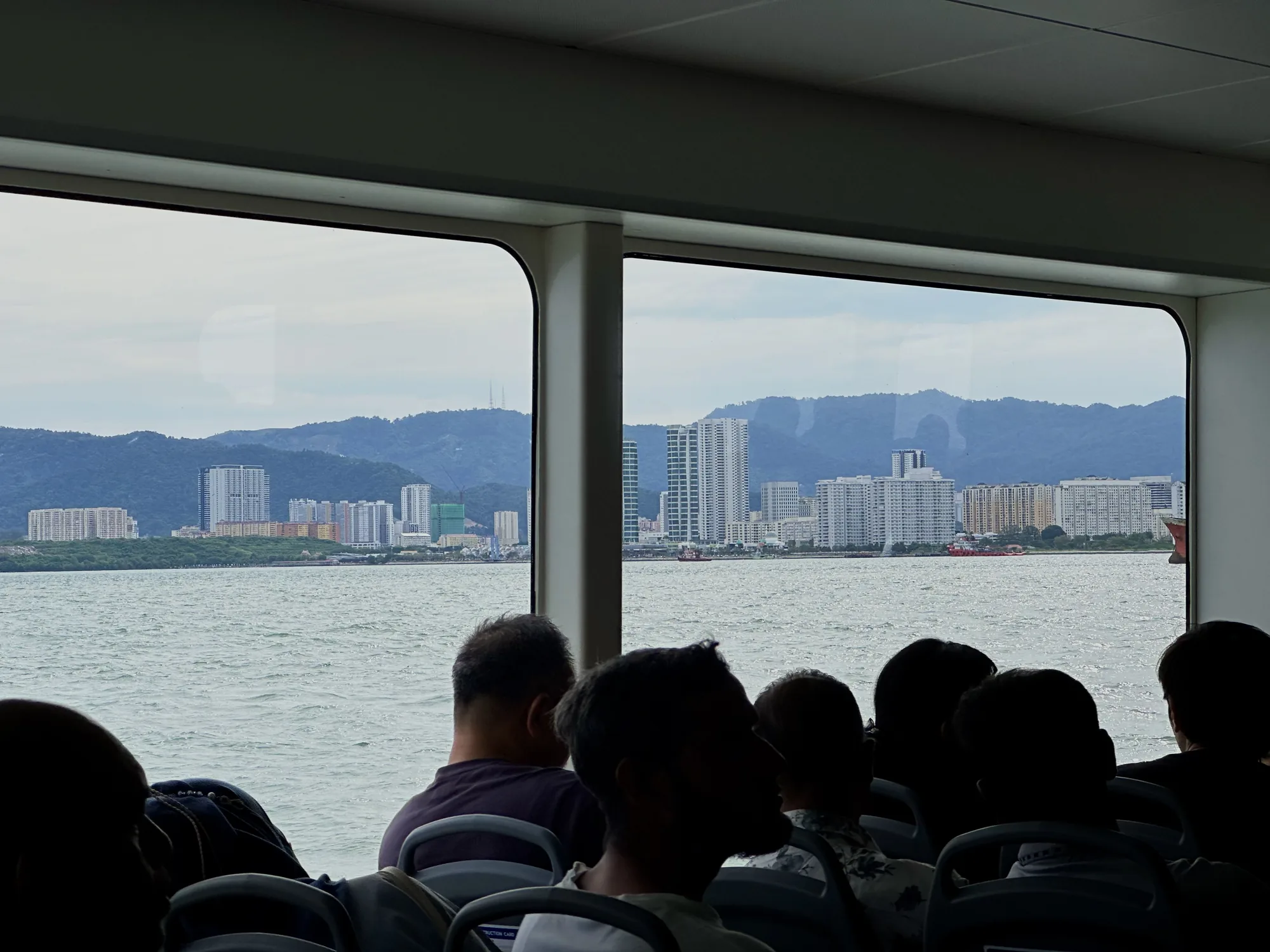 写真は、マレーシアのペナンで撮影されたもので、フェリーの窓から見える風景を捉えています。窓の外には、穏やかな海が広がり、その向こうには、緑豊かな山を背景に、高層ビル群が立ち並ぶペナンの街並みが広がっています。フェリーの中には、座席に座る乗客たちの後ろ姿が見え、彼らもまた、窓の外の景色を眺めているようです。落ち着いた雰囲気で、ゆったりとした時間が流れているように感じられます。