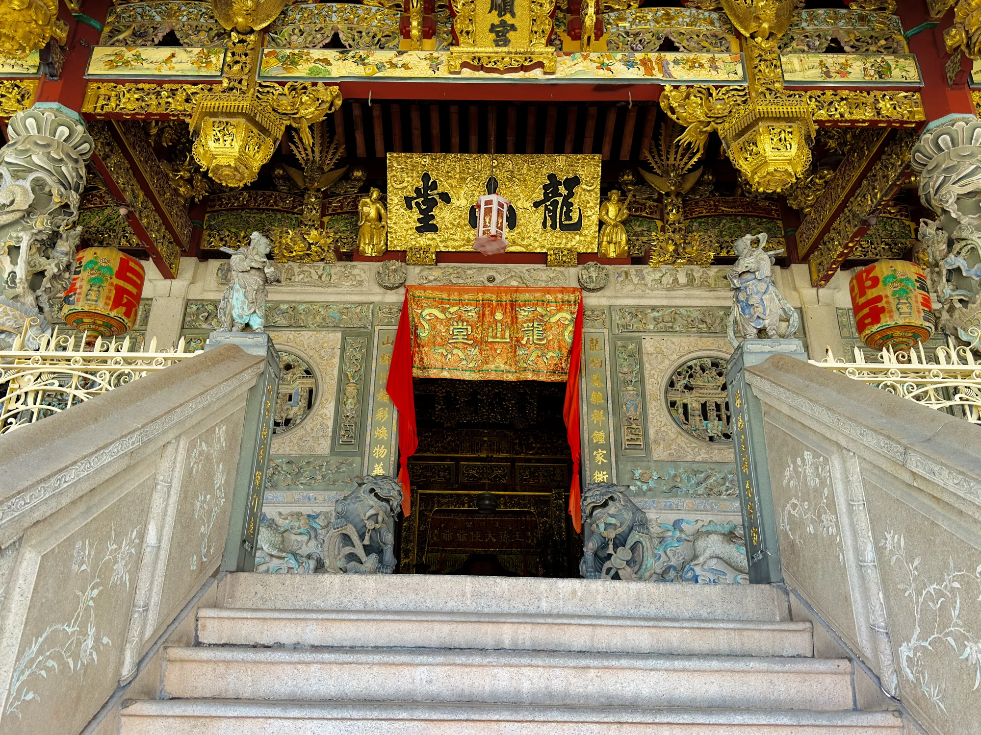 この写真はマレーシアのペナンで撮影された寺院の入り口を捉えています。石造りの階段が正面にあり、左右には手すりがあります。手すりには白い植物の装飾が施されています。階段の上には石の獅子が左右に配置され、入り口を守っているようです。入り口の上部には、金色の装飾がふんだんに施されており、豪華絢爛な雰囲気を醸し出しています。中国語の文字が書かれた金色の額や、精巧な彫刻が施された装飾品が見られます。赤色の布が入り口を飾っており、お祝いの雰囲気も感じられます。全体的に、寺院の歴史と文化を感じさせる、印象的な写真です。階段から見上げるような構図で、寺院の荘厳さを強調しています。