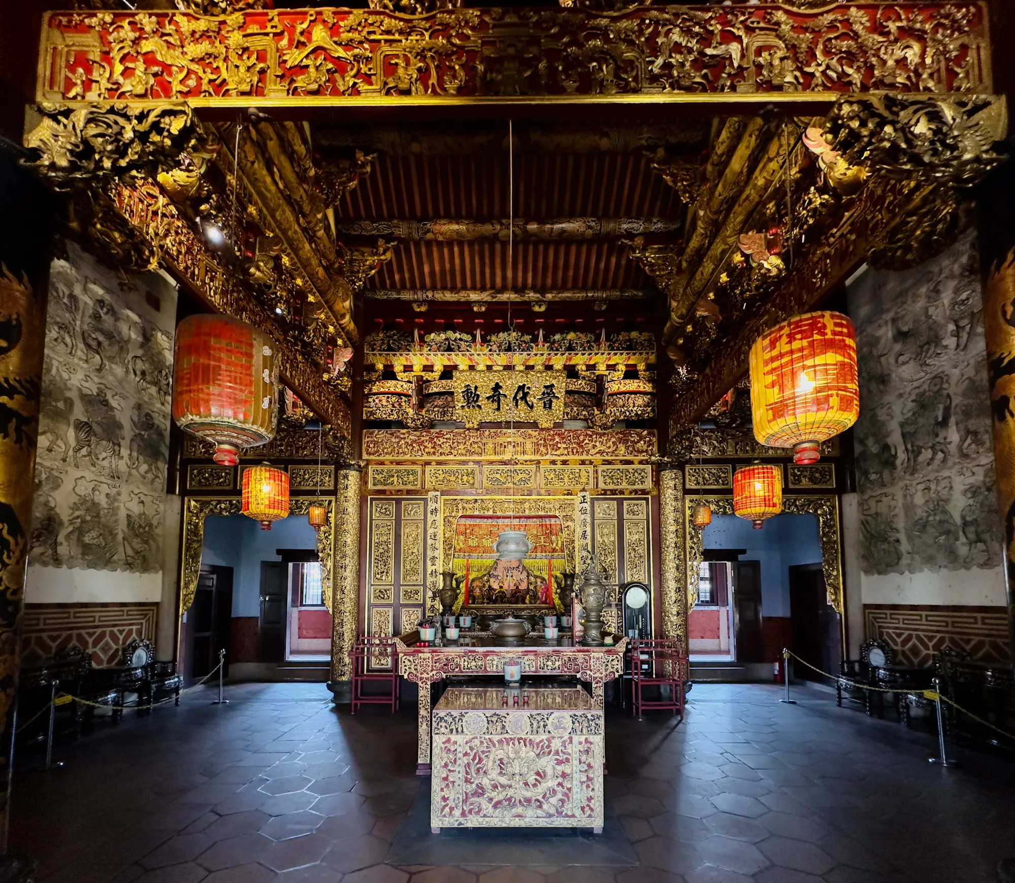 マレーシア、ペナンのある中華寺院の内部を撮影した写真です。正面には、金色の彫刻が施された祭壇があり、お供え物や香炉が置かれています。祭壇の上には「動寿代晉」と書かれた額が飾られています。左右には赤い提灯が吊り下げられており、柔らかな光を放っています。壁には水墨画のようなものが描かれています。天井は赤と金で装飾され、複雑な彫刻が施されています。床は石畳で、磨き上げられています。寺院全体は、静かで荘厳な雰囲気に包まれています。