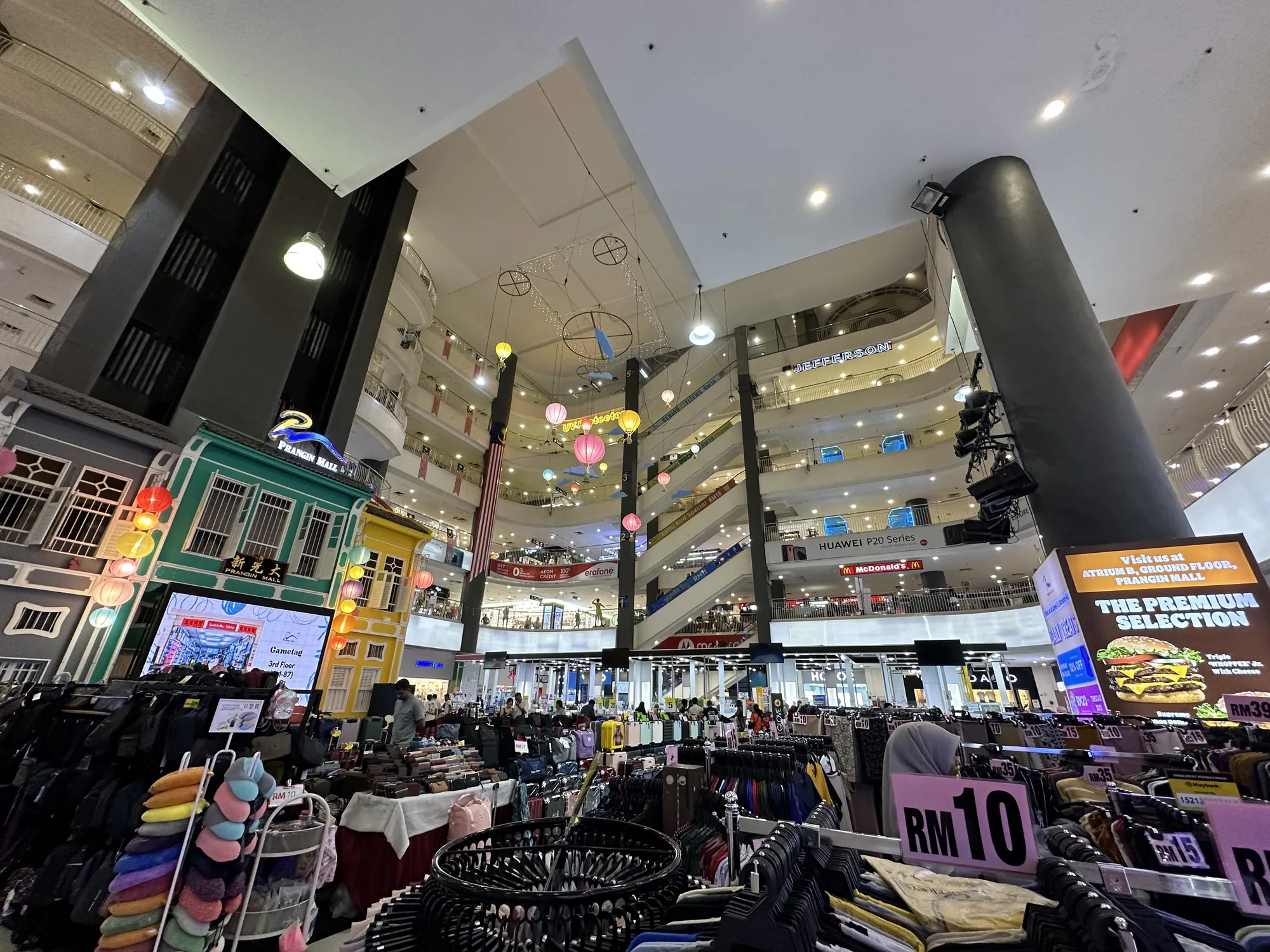 写真はマレーシアのペナンにあるショッピングモール、「Prangin Mall」の内部を捉えたものです。広角レンズで撮影されており、複数のフロアが吹き抜けになっている様子がよくわかります。天井からは装飾が吊り下げられ、各フロアには店舗が見えます。奥にはマレーシアの国旗が飾られています。  手前には衣料品などの商品が陳列された販売ブースがあり、買い物客の姿も見えます。看板やポスターも写っており、「RM 10」という価格表示が見られます。また、壁にはPrangin Mallの外観を模したカラフルな建物が装飾として設置され、ディスプレイには「Gametag」という文字が見えます。全体的に活気があり、賑やかなショッピングモールの雰囲気が伝わってきます。