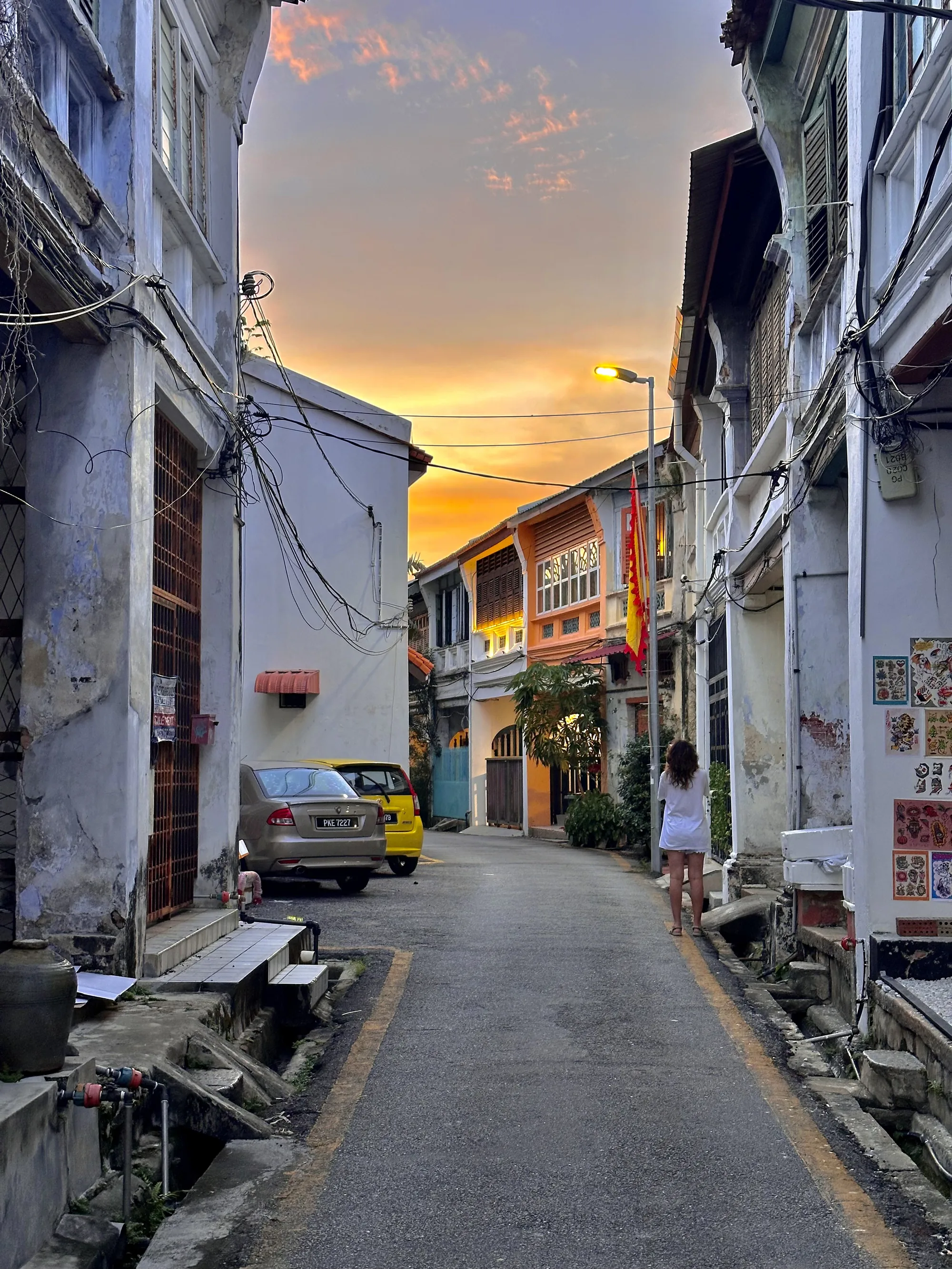 マレーシアのペナンで撮影された写真です。細い路地の奥には、夕焼け空が広がっており、オレンジ色とピンク色が混ざり合った美しいグラデーションを見せています。路地の両側には、古びた建物が立ち並び、歴史を感じさせる佇まいです。建物の壁は所々剥がれ、配線がむき出しになっている部分もありますが、それがかえって独特の風情を醸し出しています。路地の奥には黄色とベージュの車が停まっており、生活感を感じさせます。右手には、白いワンピースを着た女性が立っており、夕焼け空を眺めているようです。路地には排水溝があり、生活排水が流れているようです。壁にはステッカーが貼られており、地元の人々の日常が垣間見えます。全体的にノスタルジックで、どこか懐かしい雰囲気が漂う写真です。