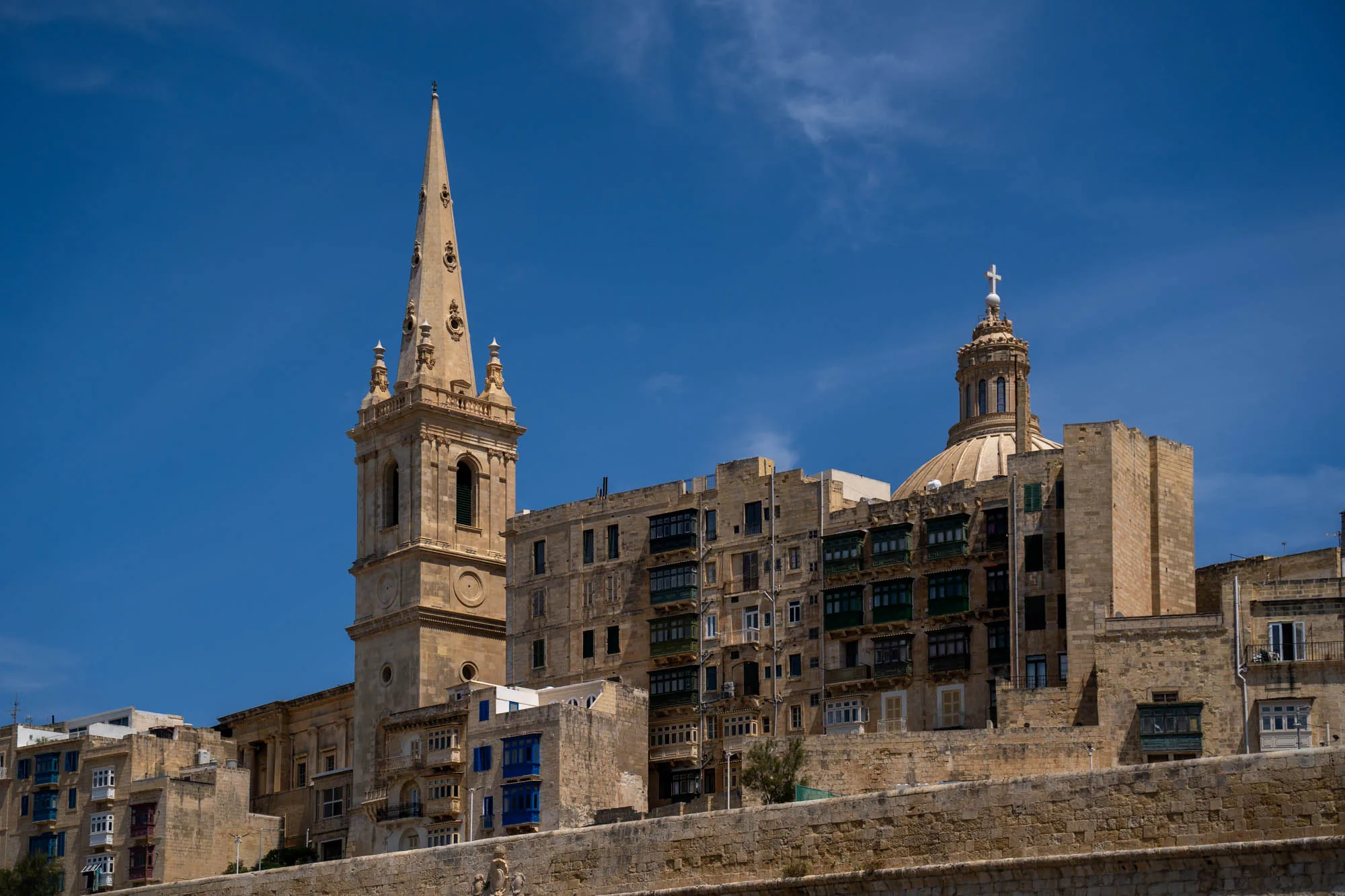 写真は、マルタの街並みを捉えたものです。青い空を背景に、ベージュ色の石造りの建物が立ち並んでいます。特に目を引くのは、左側にそびえ立つ高い尖塔を持つ教会と、右側のドーム型の屋根を持つ建物です。建物にはバルコニーが連なり、歴史を感じさせる雰囲気があります。手前の石垣が建物を支えているように見えます。全体的に明るい日差しが当たり、地中海の温暖な気候が感じられます。