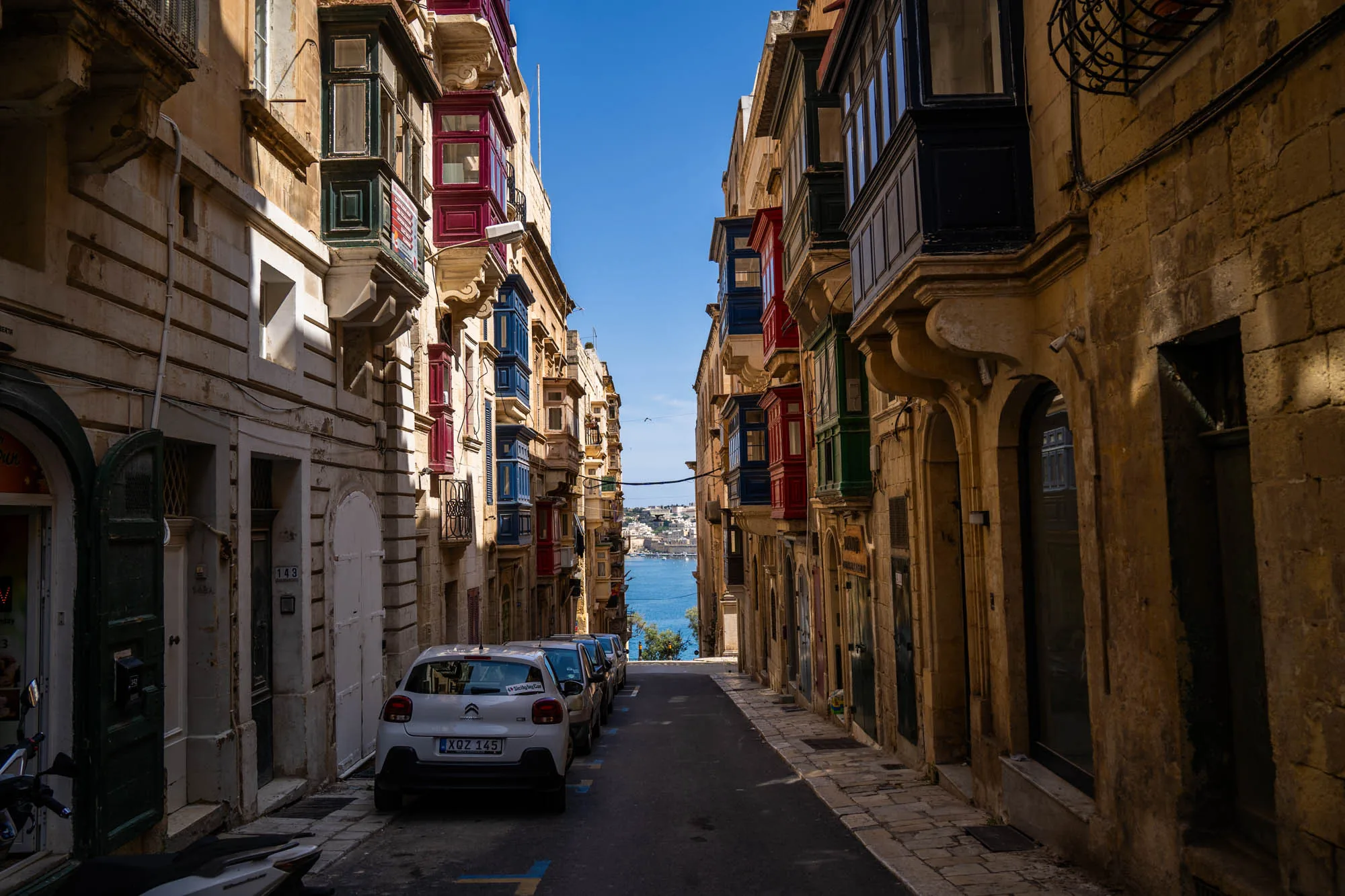 写真は、マルタの伝統的な建築様式が残る街並みを捉えたものです。石造りの建物が両側に立ち並び、その壁にはカラフルな出窓（バルコニー）が並んでいます。出窓の色は、赤、青、緑など様々で、建物の外観に彩りを添えています。路地には数台の車が駐車されており、その奥には青い海が見えます。空は晴れ渡り、遠くの島影がかすかに見えます。全体的に、歴史的な趣と地中海のリゾート地らしい明るさが感じられる写真です。建物の石の色合いや、出窓の鮮やかな色合いが美しく、視覚的に楽しめる一枚です。