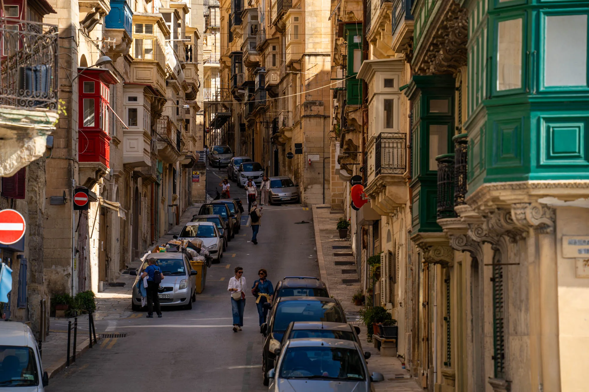 写真は、マルタの不明な場所で撮影された街並みを捉えています。狭い通りが奥へと延び、両側には特徴的な出窓を持つ古い建物が立ち並んでいます。これらの出窓は、赤、青、緑など様々な色で彩られ、それぞれ異なるデザインが施されています。建物は石造りで、経年変化による風合いが感じられます。通りには車が並んで駐車しており、数人の人々が歩いています。上り坂になっているため、奥に進むほど道幅が狭くなっているように見えます。画面左手には通行止めの標識が設置されています。全体的に、マルタの歴史的な魅力と日常生活が共存している様子が伝わってくる写真です。