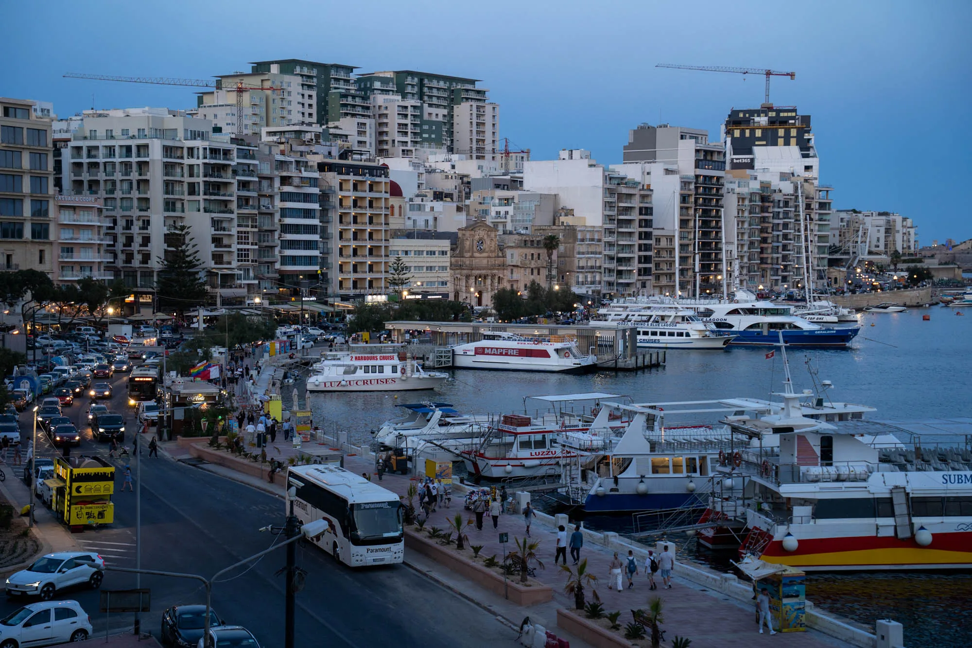 写真はマルタの港町を夕暮れ時に撮影したものです。手前には道路があり、車やバスが往来しています。道路沿いには遊歩道があり、人々が散歩を楽しんでいます。遊歩道の先は港になっており、様々な種類のボートやヨットが停泊しています。港の奥には、白い建物が密集した市街地が広がっており、建物の上には建設クレーンが見えます。空は薄暗く、夕暮れ時特有の落ち着いた雰囲気が漂っています。建物には明かりが灯り始め、港に停泊しているボートもライトアップされており、夜の始まりを感じさせます。写真全体として、マルタの港町の賑やかさと美しい景観が表現されています。遊歩道沿いにはカフェやレストランがあり、観光客や地元の人々で賑わっています。港には観光客向けのクルーズ船も停泊しており、マルタが観光地として栄えている様子が伺えます。