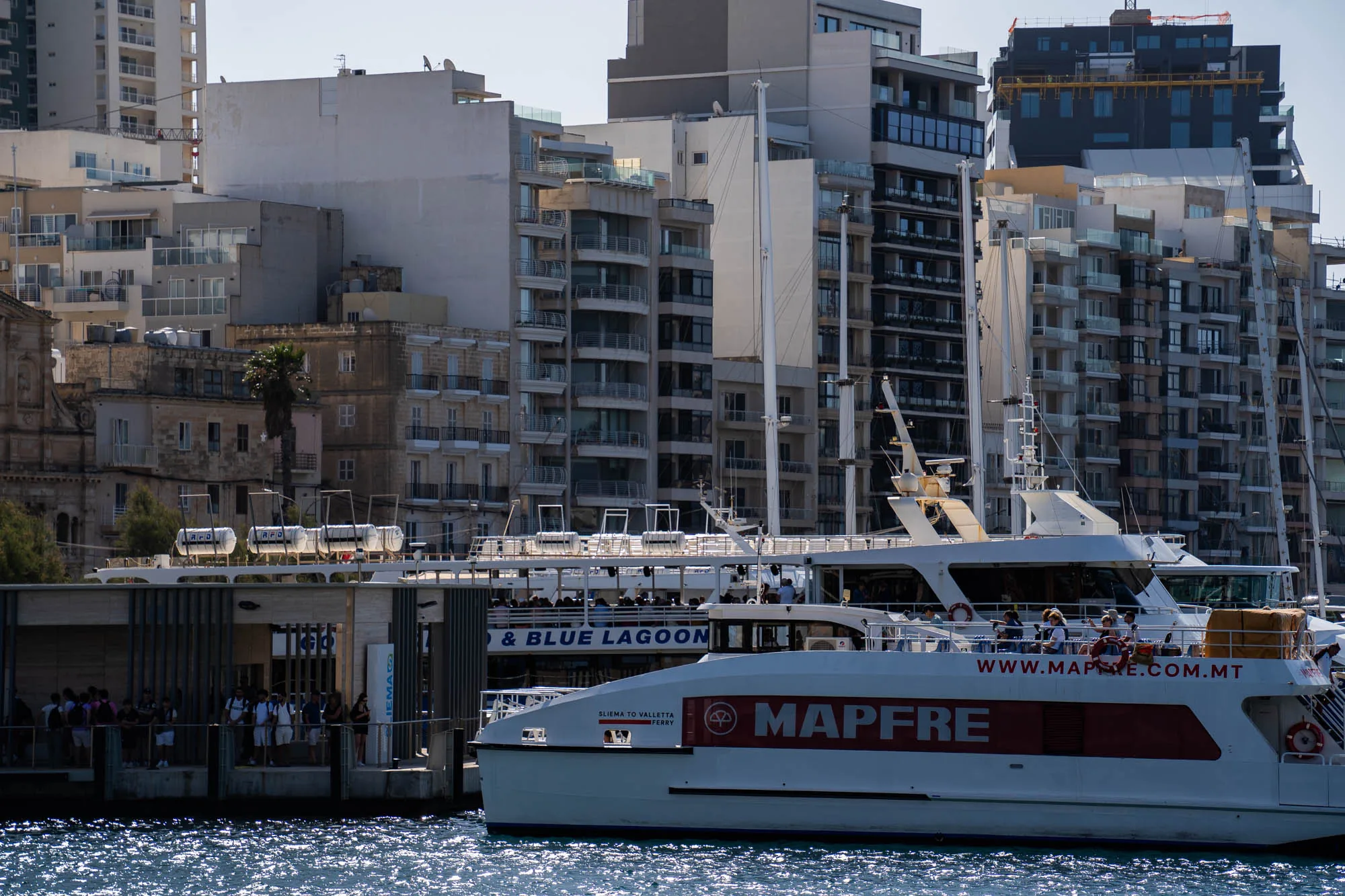 写真は、マルタの港の風景を捉えたものです。手前には、輝く水面が見え、その上に白い船が停泊しています。船体には大きく「MAPFRE」と書かれており、人々が乗船している様子が伺えます。船は「SLIEMA TO VALLETTA FERRY」と表示されており、スリーマからバレッタへのフェリーのようです。背後には、様々な高さやデザインの建物が立ち並び、都市の景観を形成しています。古い石造りの建物から、近代的な高層マンションまで、多様な建築様式が混在しており、マルタの歴史と発展を感じさせます。港には多くの人々がおり、賑わいを見せています。全体的に明るい日差しが降り注ぎ、穏やかな雰囲気です。