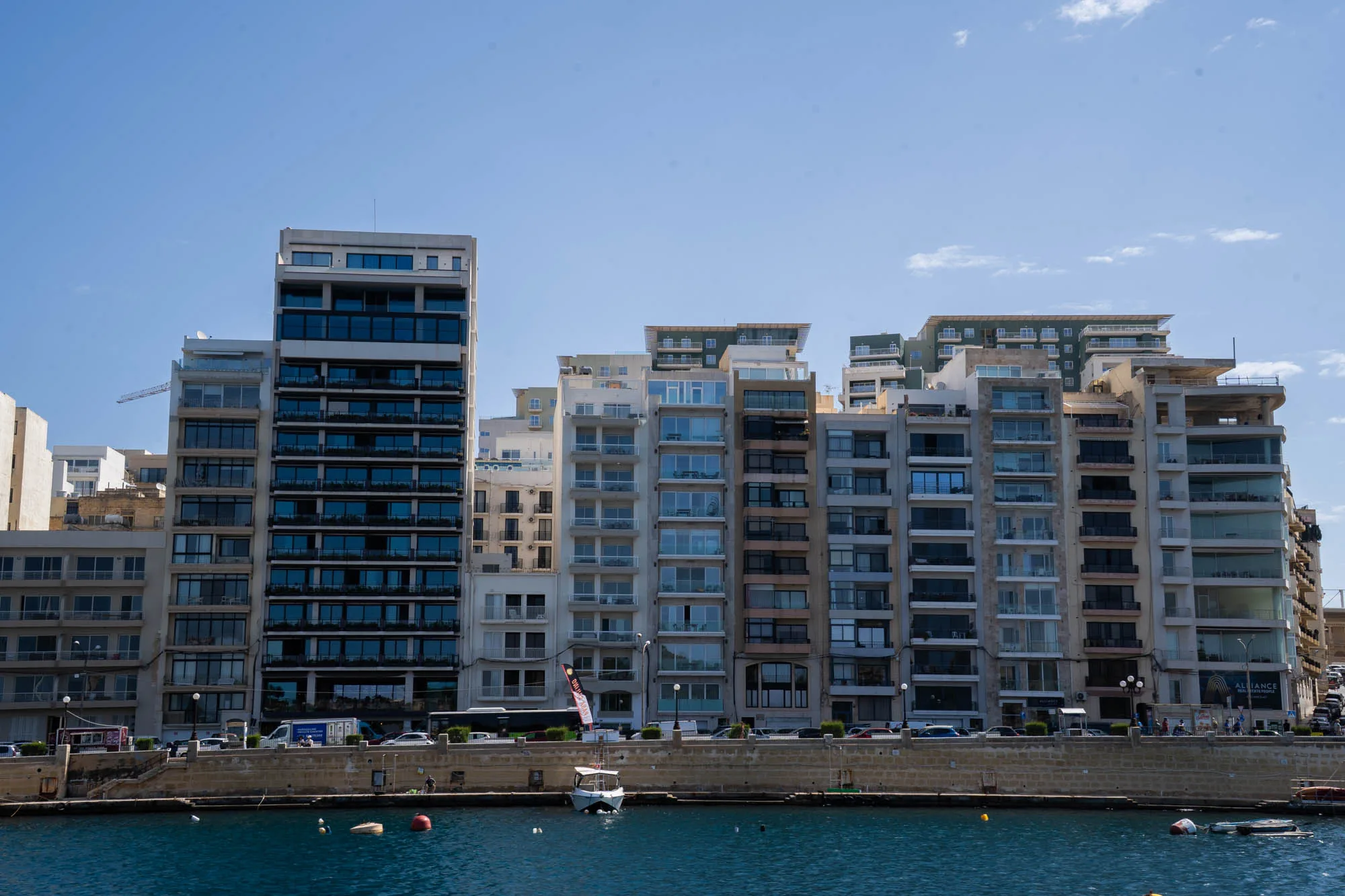 写真はマルタの海岸沿いの風景を捉えています。画面奥には、様々な高さとデザインの建物が立ち並んでいます。建物は主に白、グレー、ベージュなどの明るい色調で統一されており、バルコニーや大きな窓が特徴的です。手前には青く穏やかな海が広がり、小さなボートが停泊しています。海岸沿いの道路には、車や人が行き交う様子が見られます。全体的に明るい光に照らされており、穏やかで開放的な雰囲気が漂っています。