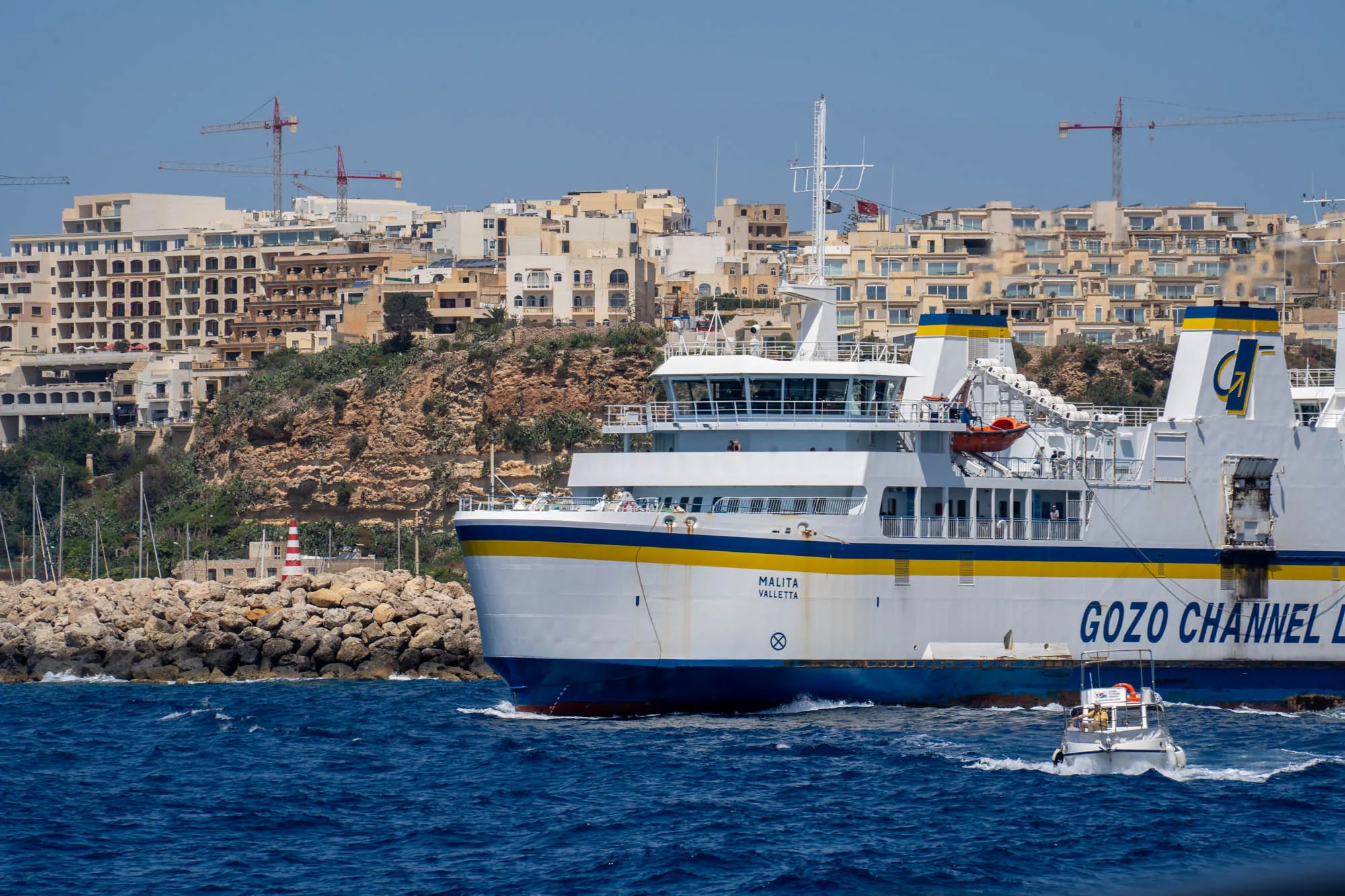 写真には、マルタの海岸線と、海を航行するフェリーが写っています。フェリーは白と青と黄色のストライプで彩られ、「MALITA VALLETTA」と船体に書かれています。船尾には「GOZO CHANNEL LINE」と書かれており、ゴゾ島への航路を運航していることがわかります。フェリーの近くには小型のモーターボートが航行しています。  背景には、崖の上に建てられた建物群が見えます。これらの建物は主にクリーム色で、地中海らしい風景を作り出しています。また、建設中のクレーンも複数見え、開発が進んでいる様子が伺えます。写真全体の色調は明るく、青い空と海が爽やかな印象を与えています。港を守る赤と白の灯台も見えます。