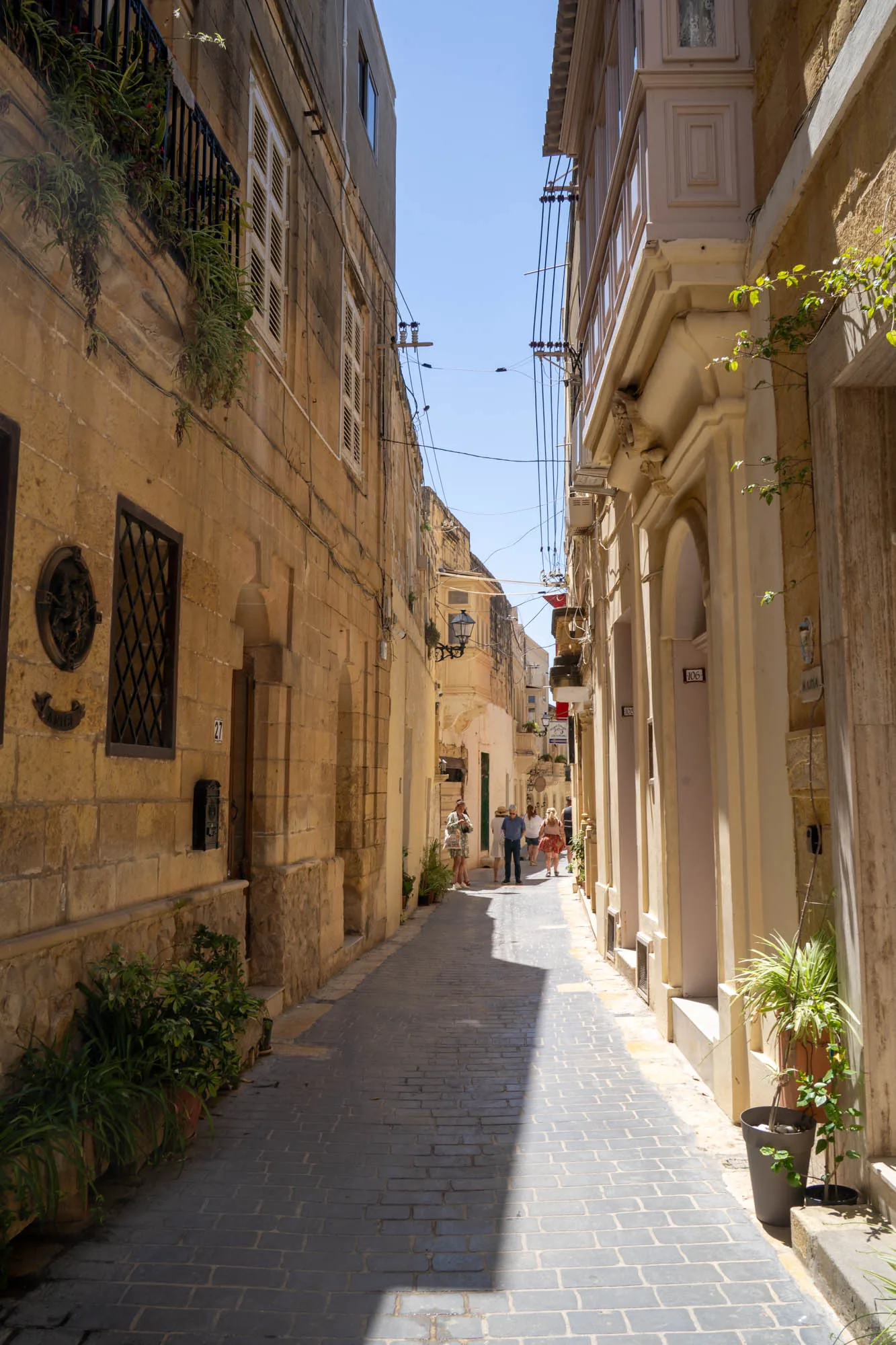 写真は、マルタの不明な場所で撮影された路地裏の風景です。道の両側には歴史を感じさせる石造りの建物が立ち並び、狭い空には青空が広がっています。建物はベージュや黄色の壁で、古い趣があります。一部の建物には、伝統的な木製のバルコニーや格子窓が見られます。建物の壁には、プランターに入った植物が飾られており、生活感と彩りを添えています。路面は石畳で、日陰と日向のコントラストが印象的です。路地の奥には数人の観光客らしき人物が歩いており、観光地の雰囲気を醸し出しています。電線が空中に張り巡らされているのも、ヨーロッパの古い街並みによく見られる光景です。全体的に、マルタの歴史的な街並みの魅力と、ゆったりとした時間が流れる雰囲気が伝わってくる写真です。