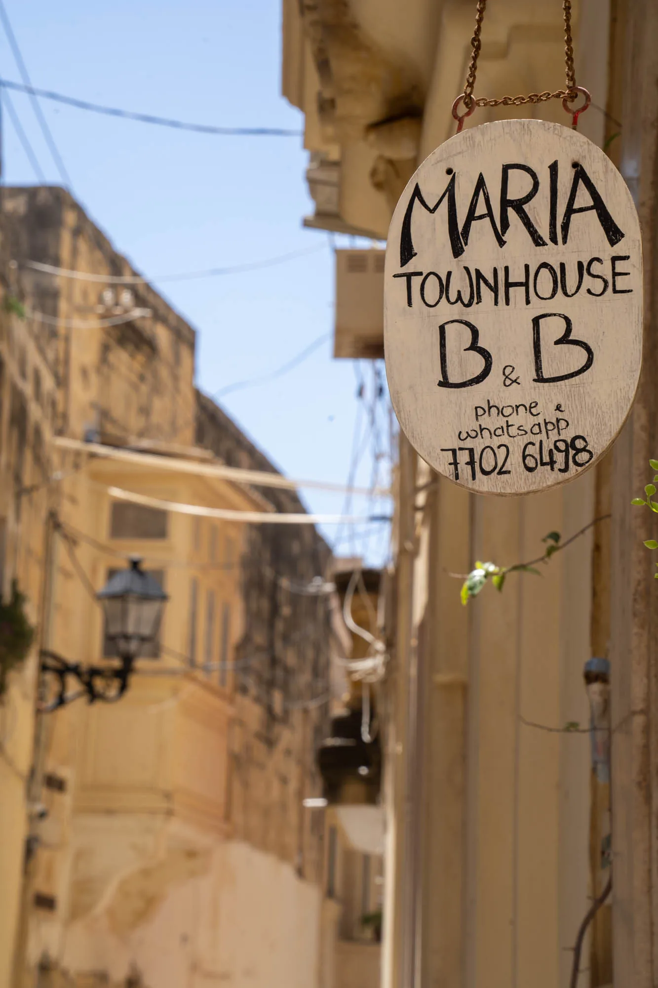 マルタの狭い路地に面した場所で撮影された写真です。手前には、「MARIA TOWNHOUSE B&B」と書かれた白い木製の看板が、鎖で吊り下げられています。看板には電話番号とWhatsAppの連絡先も記載されています。背景には、薄いベージュ色の石造りの建物が並ぶ路地の風景が広がっています。路地は狭く、建物は古く、歴史を感じさせる雰囲気です。路地の奥には街灯が見えます。空は青く、日差しが強く、マルタの夏の日の雰囲気を伝えています。