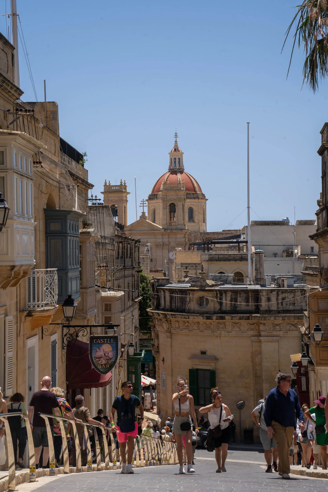 写真はマルタの街並みを捉えたものです。緩やかな坂道の石畳の道があり、その両脇には歴史を感じさせる建物が立ち並んでいます。建物は主にベージュ色で、特徴的な出窓（バルコニー）や装飾が施されています。遠景には赤茶色のドームを持つ教会が見え、街のランドマークとなっています。画面手前には観光客と思われる人々が歩いており、マルタの街並みを散策している様子が伺えます。「CASTLE BAR」という看板も確認でき、観光地であることがわかります。明るい日差しが降り注ぎ、穏やかな雰囲気です。