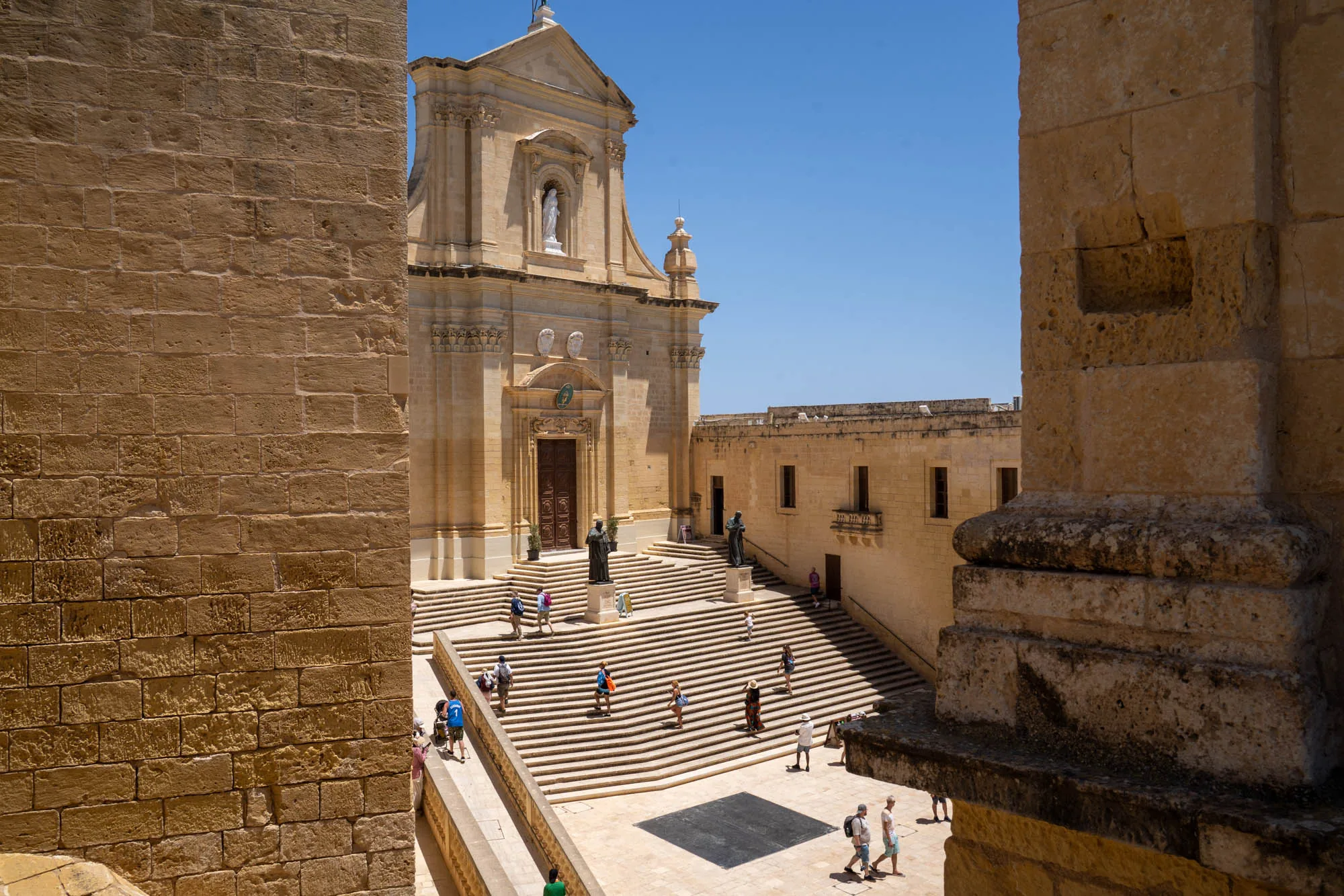 写真はマルタで撮影された、明るい日中の風景です。石造りの建物の一部をフレームのように左右に配置し、その間から、大きな教会と、階段を上り下りする人々が見えます。教会は淡い黄色の石でできており、正面には聖母像が安置されています。階段は広くて段数が多く、大勢の観光客が歩いています。建物の石の色と、青空の色がコントラストをなし、暖かく穏やかな雰囲気を作り出しています。左右の石壁の質感や陰影が、写真に奥行きと立体感を与えています。