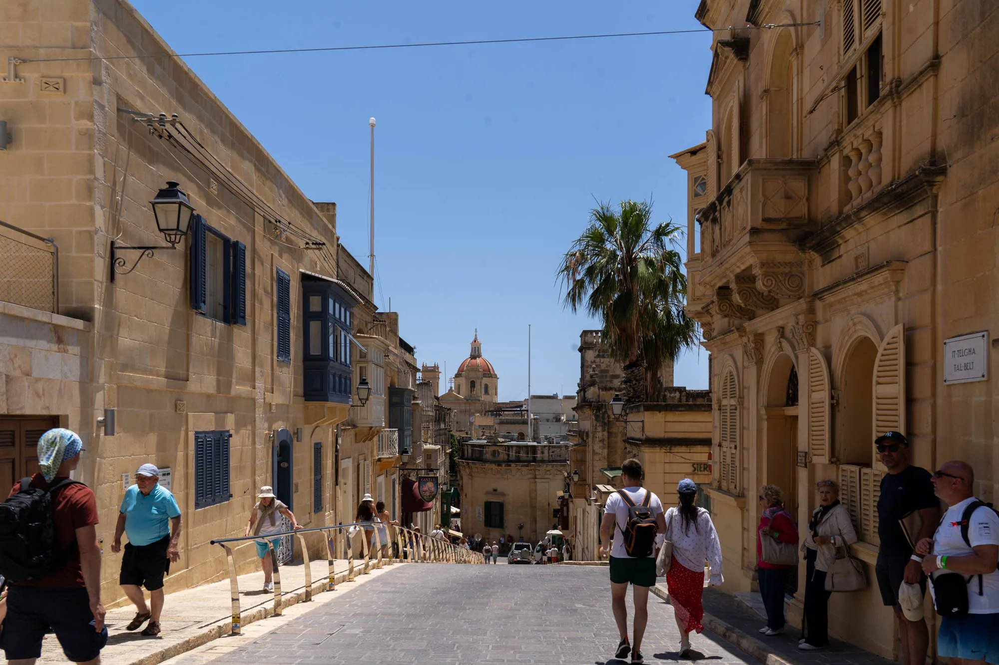 写真はマルタの不明な場所で撮影された街並みの様子を捉えています。石畳の坂道が奥へと続き、両側にはハニーカラーの石造りの建物が立ち並んでいます。建物には青い鎧戸やバルコニーが特徴的で、地中海らしい雰囲気を醸し出しています。空は晴れ渡っており、坂道の先には教会のドームが見えます。道には観光客らしき人々が歩いており、活気のある街の様子が伝わってきます。画面右側には「IT-TELGHA TAL-BELT」と書かれた白い標識が確認できます。