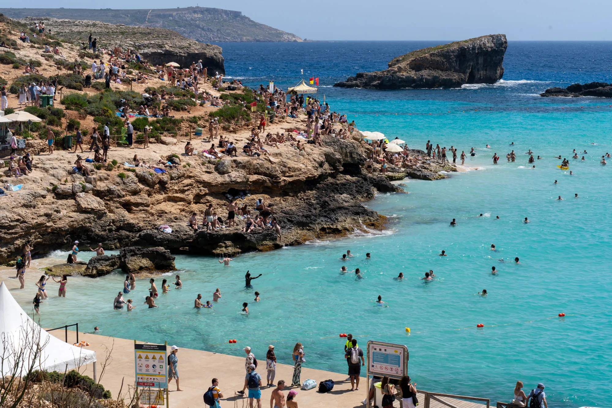 写真は、マルタの海岸の風景を捉えたものです。ターコイズブルーの美しい海が印象的で、多くの人々が海水浴を楽しんでいます。海岸線は岩場で、日当たりの良い岩場には多くの人々が座って日光浴をしたり、談笑したりしています。海の中にもたくさんの人がいて、泳いだり、水遊びをしたりしています。背景には、海に突き出した岩山があり、その上に緑が生い茂っています。空は晴れていて、全体的に明るく、賑やかな夏の休暇の雰囲気を感じさせます。写真の下部には、木製のデッキがあり、人々が歩いています。海岸沿いには、安全に関する注意書きの看板も見られます。全体的に、マルタの美しい自然と、人々が休暇を楽しんでいる様子が伝わってくる写真です。