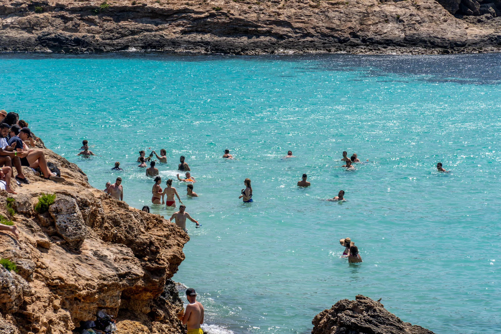 写真には、マルタの美しい海岸線が写っています。特に目を引くのは、透き通ったターコイズブルーの海です。多くの人々が水着姿で海に入り、泳いだり、日光浴を楽しんだりしています。海岸線は岩場で、一部の人々は岩の上に座ってリラックスしています。海の向こうには、岩がちな海岸線が続いており、その先には濃い青色の海が広がっています。全体的に、夏の日の活気あるビーチの雰囲気が伝わってきます。水面には太陽の光が反射し、きらきらと輝いています。