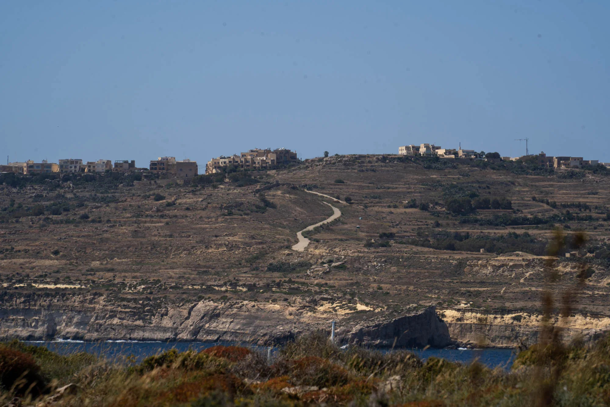 写真はマルタの乾燥した風景を捉えています。全体的に茶色やベージュの色合いが強く、乾燥した土地が広がっていることがわかります。遠景には建物が連なっており、丘の上に集落があるようです。丘の中腹には曲がりくねった道が通っており、集落へと繋がっています。中景には、崖のような地形が見られ、その下には青い海が広がっています。前景は、草や低木などが生い茂っており、少しぼやけています。空は晴れており、全体的に明るい印象です。マルタの乾燥した気候と独特の地形が伝わる風景写真です。