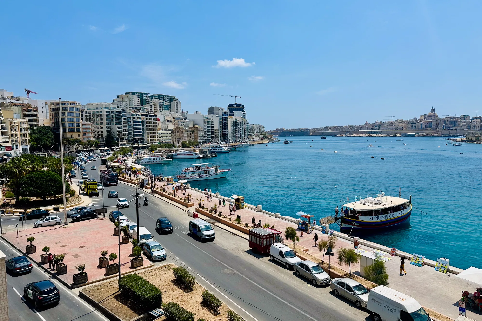 写真はマルタの都市部で撮影されたもので、港町の風景が広がっています。鮮やかな青色の海が印象的で、数多くのボートが停泊または航行しています。海岸沿いには歩道が整備されており、人々が散策を楽しんでいます。道路には車が走り、活気のある様子が伺えます。背景には、高層ビルや歴史的な建造物などが立ち並び、都市の景観を形作っています。空は晴れ渡り、わずかに雲が見えます。全体的に、マルタの美しい港町とその活気ある雰囲気を捉えた写真です。