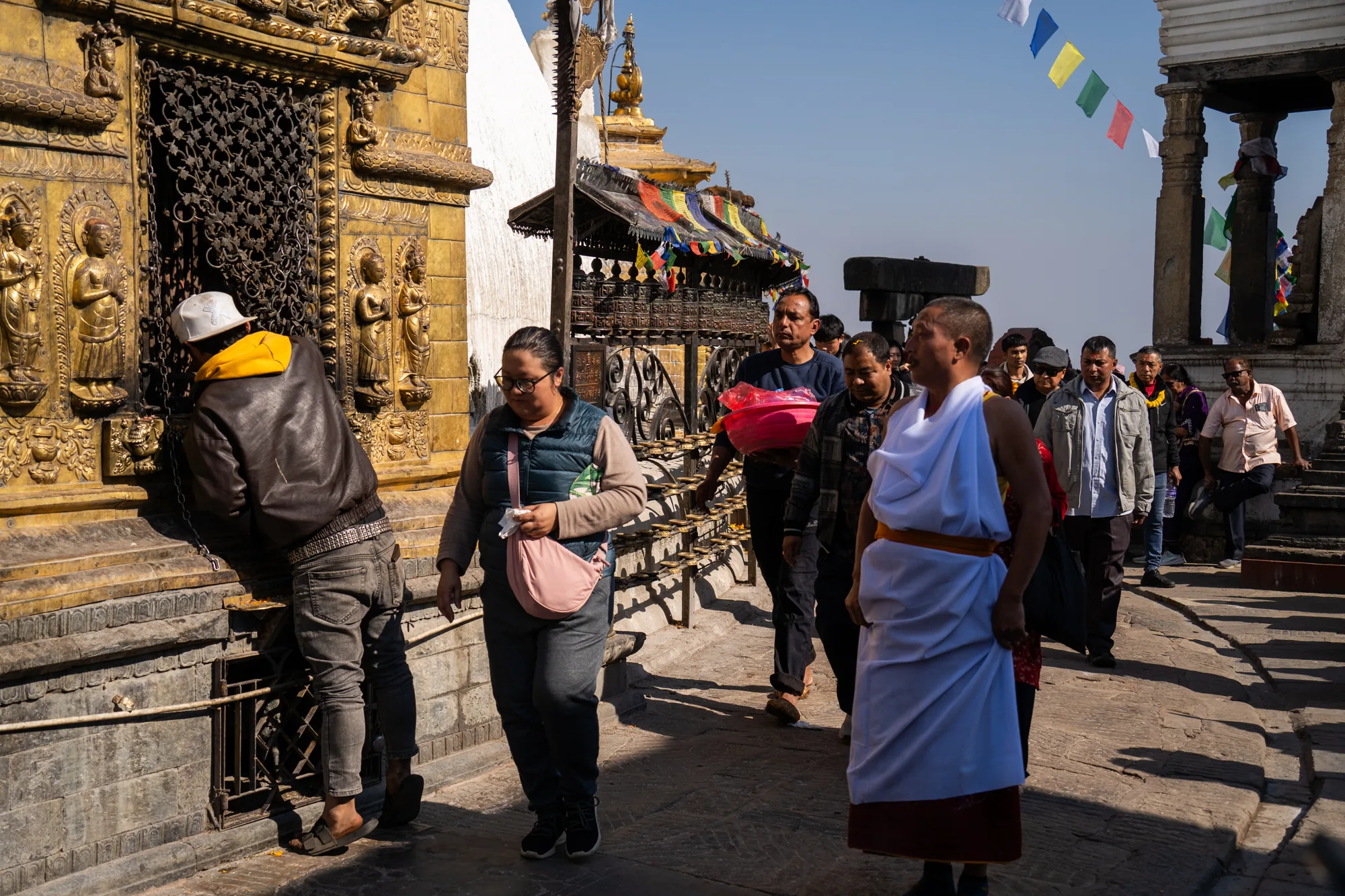 ネパールのカトマンズで撮影されたと思われる写真です。寺院と思われる場所で、参拝者や観光客など様々な人々が行き交っています。画面左側には金色の装飾が施された壁があり、格子状の窓の前に男性が立って何かを覗き込んでいます。その隣には女性が歩いており、ピンク色のバッグを持っています。  中央から右側にかけては、人々が列をなして歩いています。僧侶の服装をした人物、民族衣装をまとった人、普段着の人など様々です。奥には仏塔のような建物が見え、カラフルな旗が飾られています。空は晴れており、日差しが強く、地面には影がはっきりと映っています。全体的に活気があり、宗教的な雰囲気と人々の生活が混ざり合っている様子が伝わってきます。
