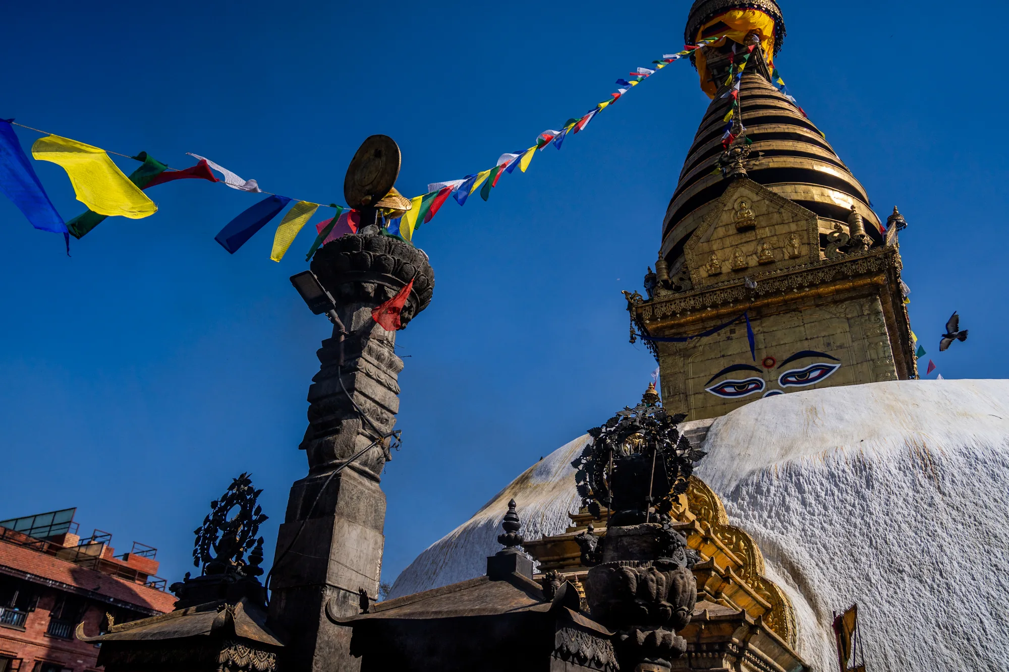 ネパールのカトマンズで撮影された写真には、仏塔が写っています。青空を背景に、白いドーム型の基壇の上にそびえ立つ金色の塔が印象的です。塔には仏陀の目が描かれており、神聖な雰囲気を醸し出しています。  写真全体に、タルチョと呼ばれる五色の祈祷旗が張り巡らされており、風に揺らめいています。これらの旗は、平和や幸福などの願いが込められています。また、石造りの構造物も写っており、歴史を感じさせる趣があります。鳥が空を飛んでいる様子も捉えられており、風景に動きを与えています。全体的に、鮮やかな色彩と歴史的な建造物が組み合わさった、カトマンズの文化的景観を捉えた写真です。