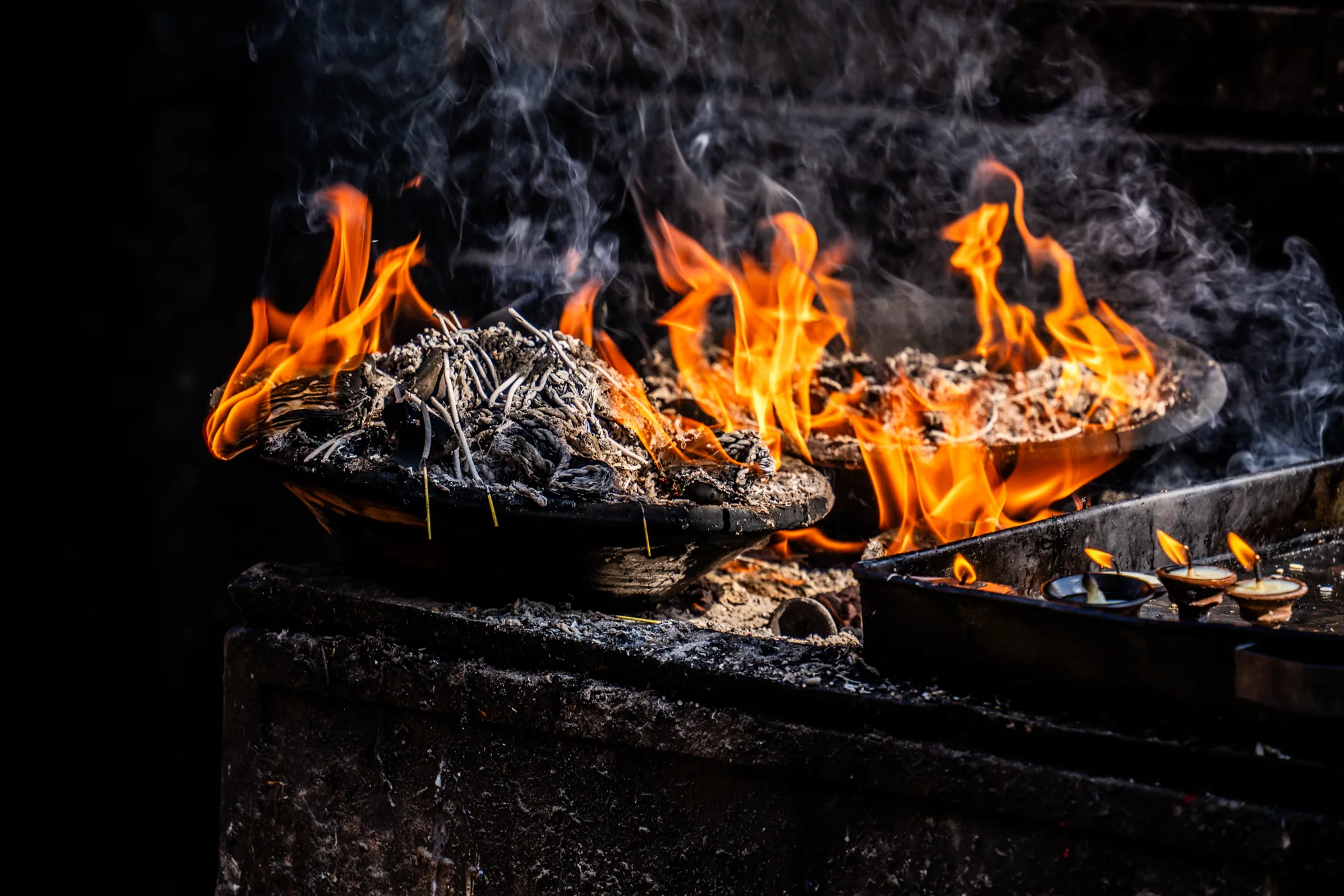 写真はネパールのカトマンズで撮影されたもので、宗教的な儀式に使われる火を捉えています。背景は暗く、焦点は燃え盛る炎と立ち上る煙に当てられています。二つの黒い器には、燃え尽きた灰と煙が立ち込める中、激しく炎が燃え盛っています。手前には、油が満たされた小さな器が並んだ金属製のトレイがあり、それぞれの器の中で小さな炎が静かに揺らめいています。全体的に神聖で神秘的な雰囲気が感じられ、宗教的な儀式や信仰を連想させます。炎と煙の躍動感が印象的な一枚です。
