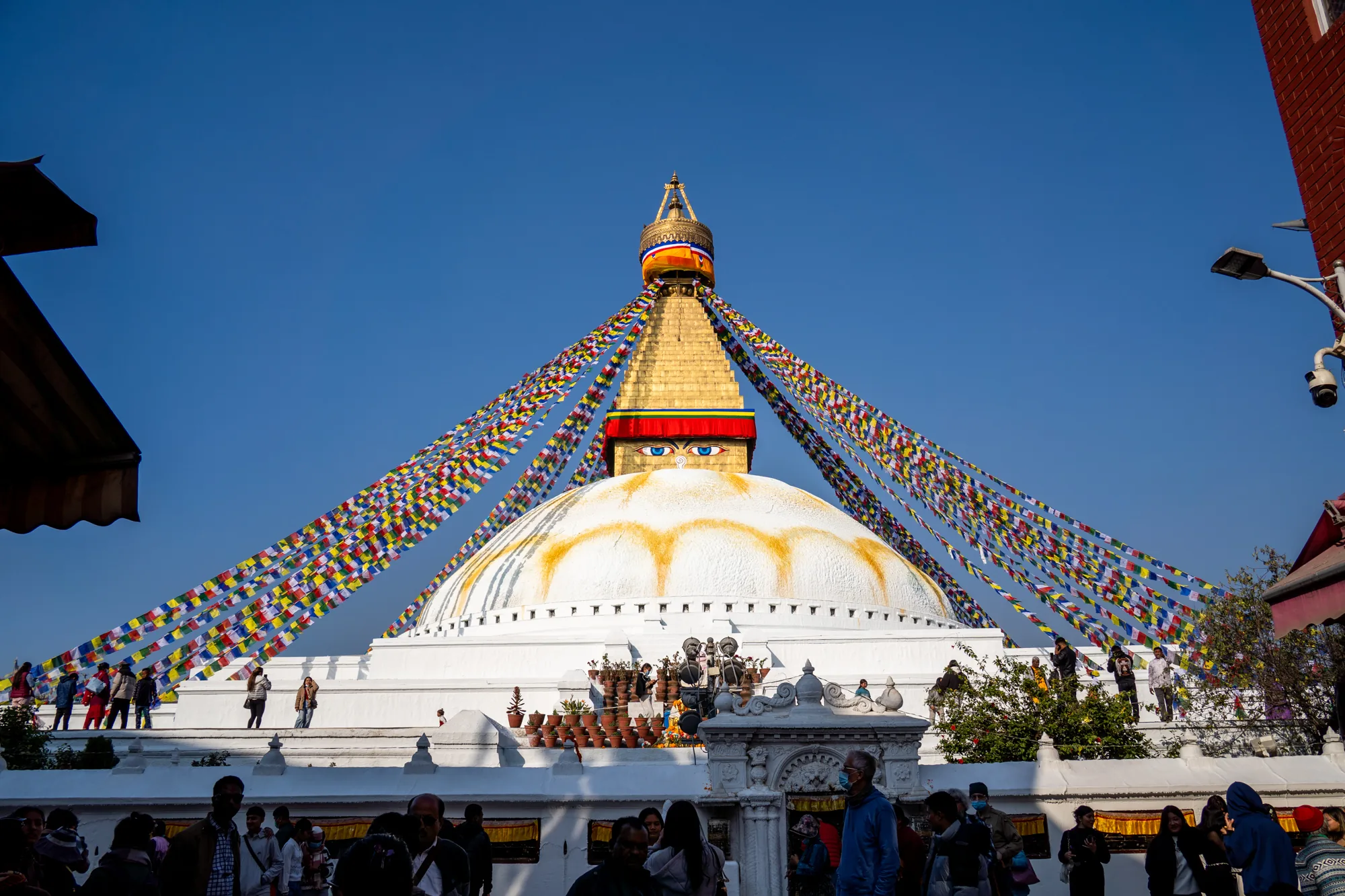 この写真はネパールのカトマンズにあるボダナート（またはボダナートストゥーパ）と呼ばれる仏塔を捉えたものです。空は晴れ渡っており、仏塔の白色と金色のコントラストが際立っています。仏塔の頂上には、金色の装飾が施された部分があり、その下には四角い構造物があり、その四方には仏陀の目が描かれています。仏塔全体からタルチョ（祈祷旗）が四方八方に張り巡らされ、青空に鮮やかな色彩を添えています。  仏塔の基部には、階段状の構造があり、人々が訪れ、写真を撮ったり、散策したりしています。仏塔の周囲には、多くの人々が集まっており、様々な服装をしています。写真の右側には建物の一部が見え、街灯や監視カメラも設置されています。全体的に、活気と静寂が共存する、宗教的な場所の雰囲気が伝わってきます。