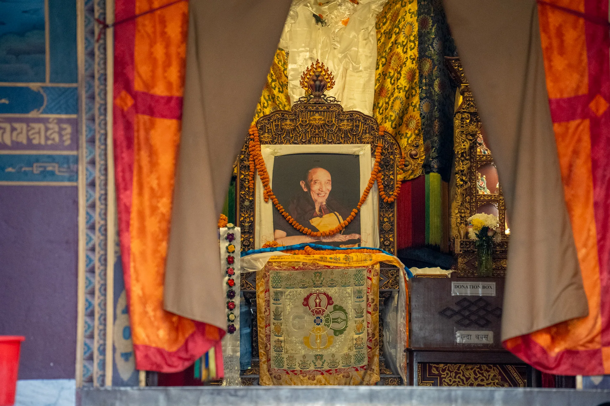 この写真はネパールのカトマンズで撮影されたもので、僧侶を祀る祭壇を捉えています。祭壇の中心には、笑顔の僧侶の肖像画が飾られており、肖像画の額縁は精巧な彫刻が施された金色の装飾で飾られています。肖像画にはマリーゴールドの花輪がかけられています。祭壇はカラフルな織物で覆われており、チベット仏教のシンボルが描かれています。祭壇の奥には、金色の装飾が施された別の祭壇が見え、仏像と花が飾られています。写真全体は、オレンジや黄色の色調が強く、神秘的で精神的な雰囲気を醸し出しています。左側には、模様の描かれた壁が見え、右側には寄付箱が置かれています。