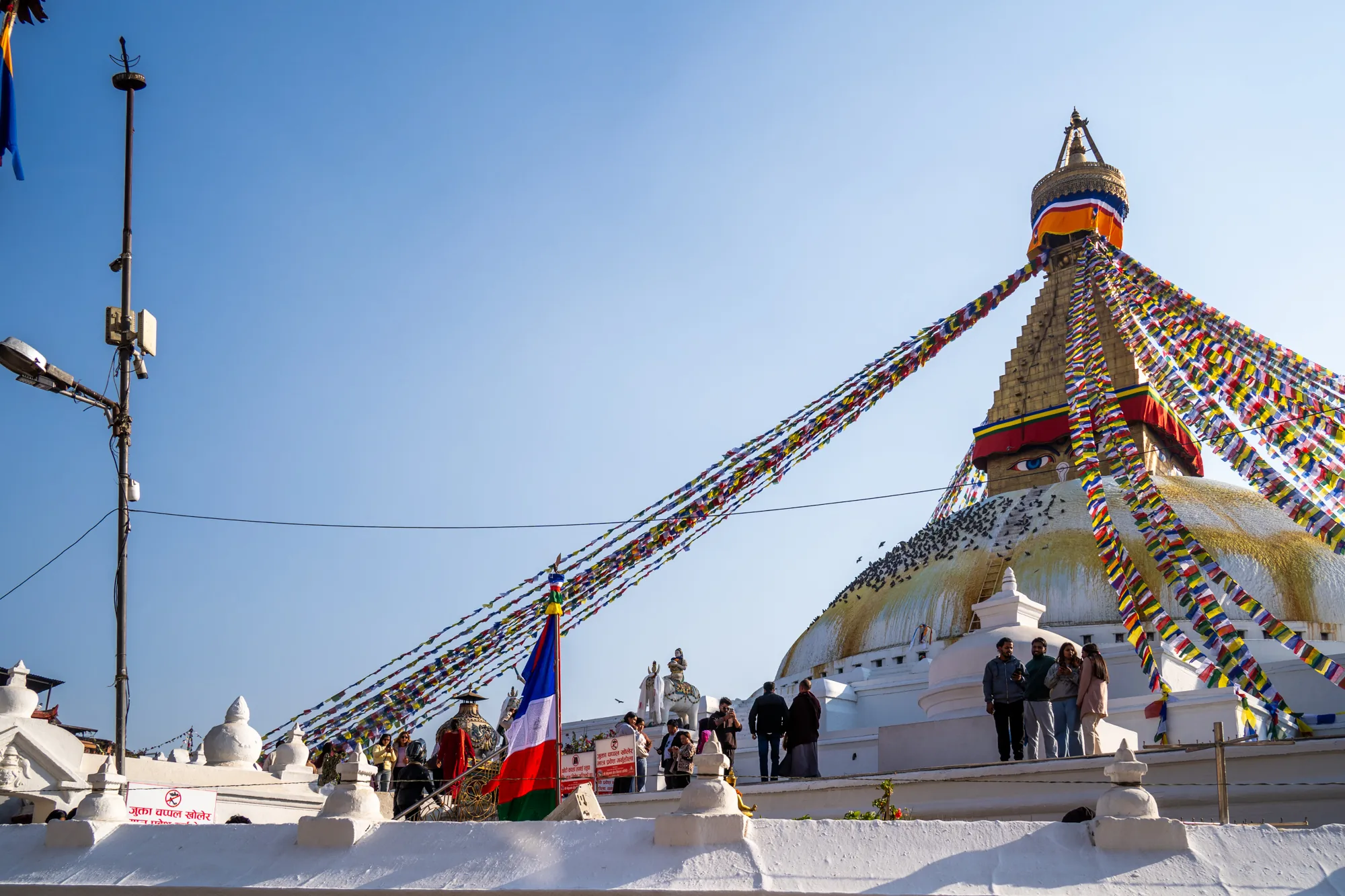 ネパールのカトマンズにあるボダナートの風景を捉えた写真です。青空を背景に、巨大な白いドーム型のストゥーパがそびえ立っています。ストゥーパの頂上には、金色の塔があり、カラフルなタルチョ（祈祷旗）が四方八方に張り巡らされています。ストゥーパの白い壁面には、たくさんの鳥が群がっています。ストゥーパの周りには、観光客と思われる人々が散策しており、ネパールの国旗も掲げられています。全体的に、晴天の下で神聖な場所を訪れる人々の活気が感じられる写真です。