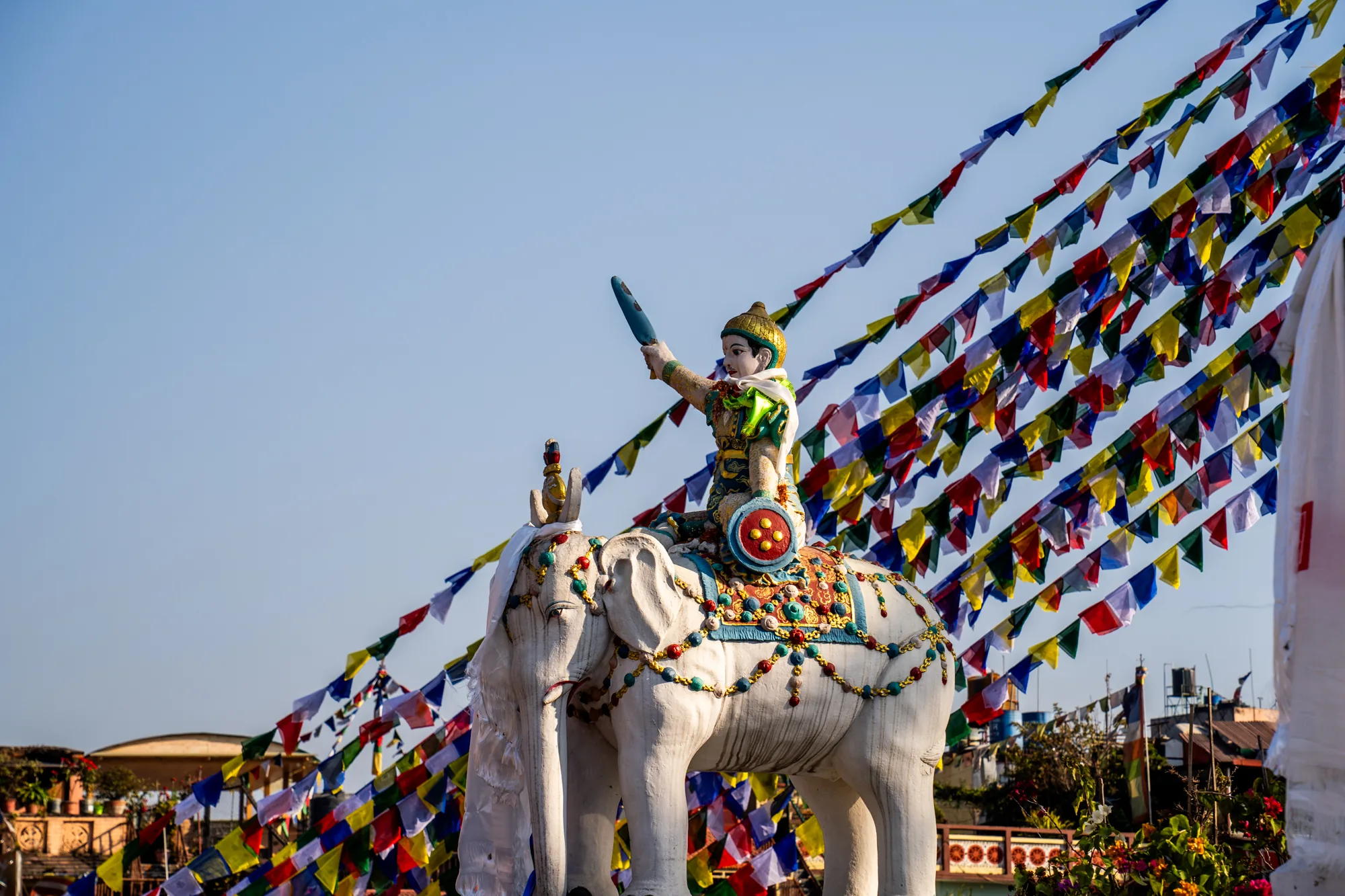 この写真はネパールのカトマンズで撮影されたものです。青空を背景に、白い象の像が大きく写っています。象の背中には、剣のようなものを持った人物の像が乗っています。象は金色の装飾やカラフルな装飾が施されており、特に背中の装飾は鮮やかです。象の周囲には、チベット仏教のタルチョ（祈祷旗）が何本も連なっており、赤、青、黄、緑、白など色とりどりの旗が風になびいている様子が写っています。タルチョは建物の屋上から張られているようです。背景には、カトマンズの建物の一部が写っており、異国情緒あふれる風景が広がっています。全体的に、文化的な要素が強く、鮮やかで活気のある雰囲気の写真です。