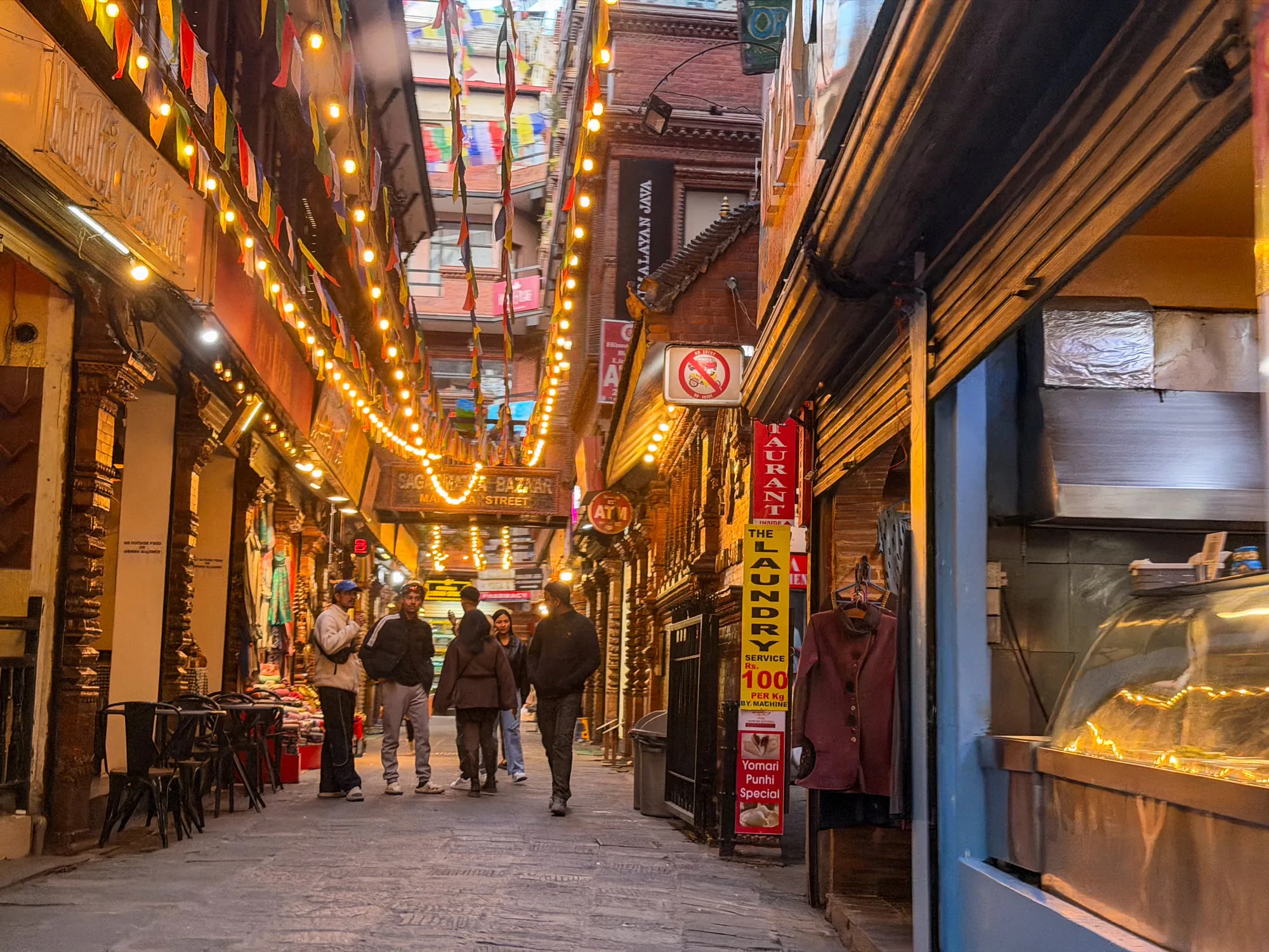 写真は、ネパールのカトマンズにある活気のある通りの風景を捉えています。石畳の道には人々が歩いており、両側にはお店が立ち並んでいます。通りの上部には、色とりどりの祈祷旗が飾られ、温かい光を放つ電球のストリングライトが吊るされています。これらの装飾が、街の賑やかさと文化的な魅力を強調しています。左側の店舗には椅子が並べられ、客を待っているようです。右側には「THE LAUNDRY」と書かれた看板があり、奥にはレストランらしき場所が見えます。全体的に、この写真はカトマンズの通りのにぎやかで魅力的な雰囲気を伝えています。