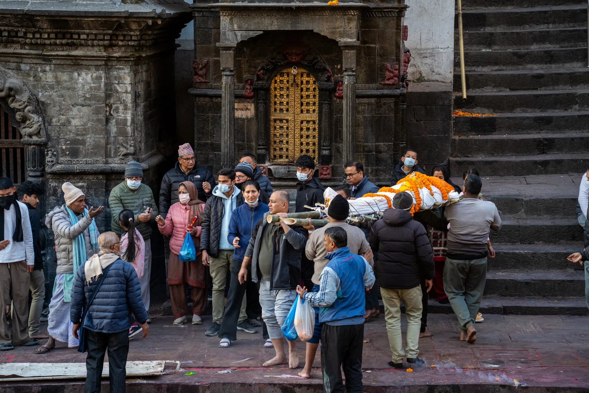 この写真はネパールのパシュパティナートで撮影されたもので、葬儀の様子が写されています。中央には、花で飾られた遺体が担架に乗せられ、運ばれています。遺体の周りには、マスクを着用した人々を含む、多くの参列者が集まっています。彼らの表情は様々で、悲しみや敬意、あるいは静かな祈りを捧げている様子が見て取れます。背景には、階段や装飾が施された古い建物が写っており、歴史的な場所であることが分かります。地面には花びらが散らばっており、葬儀の儀式の一部であったことを示唆しています。全体的に、この写真は死と哀悼、そして文化的な伝統が混ざり合った、厳粛な雰囲気を伝えています。
