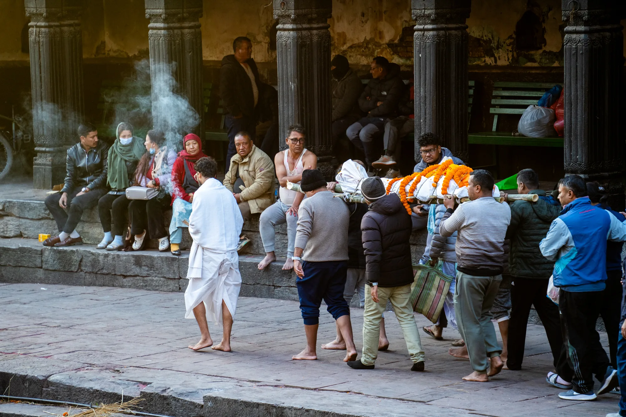 写真はネパールのパシュパティナートで撮影されたものです。石畳の広場のような場所で、手前に火葬の煙が漂っています。中央には、オレンジ色の花で飾られた白い布に包まれた棺を担ぐ人々がいます。棺を担ぐ人々の他に、白い布をまとった男性、半袖の男性、座って様子を見ている人々など様々な人たちが写っています。背景には、石造りの柱が並び、ベンチに腰掛けている人々の姿も見えます。全体的に落ち着いた色調で、神聖で厳粛な雰囲気が漂っています。生活と死が隣り合わせにあるような、独特な空気感が感じられます。