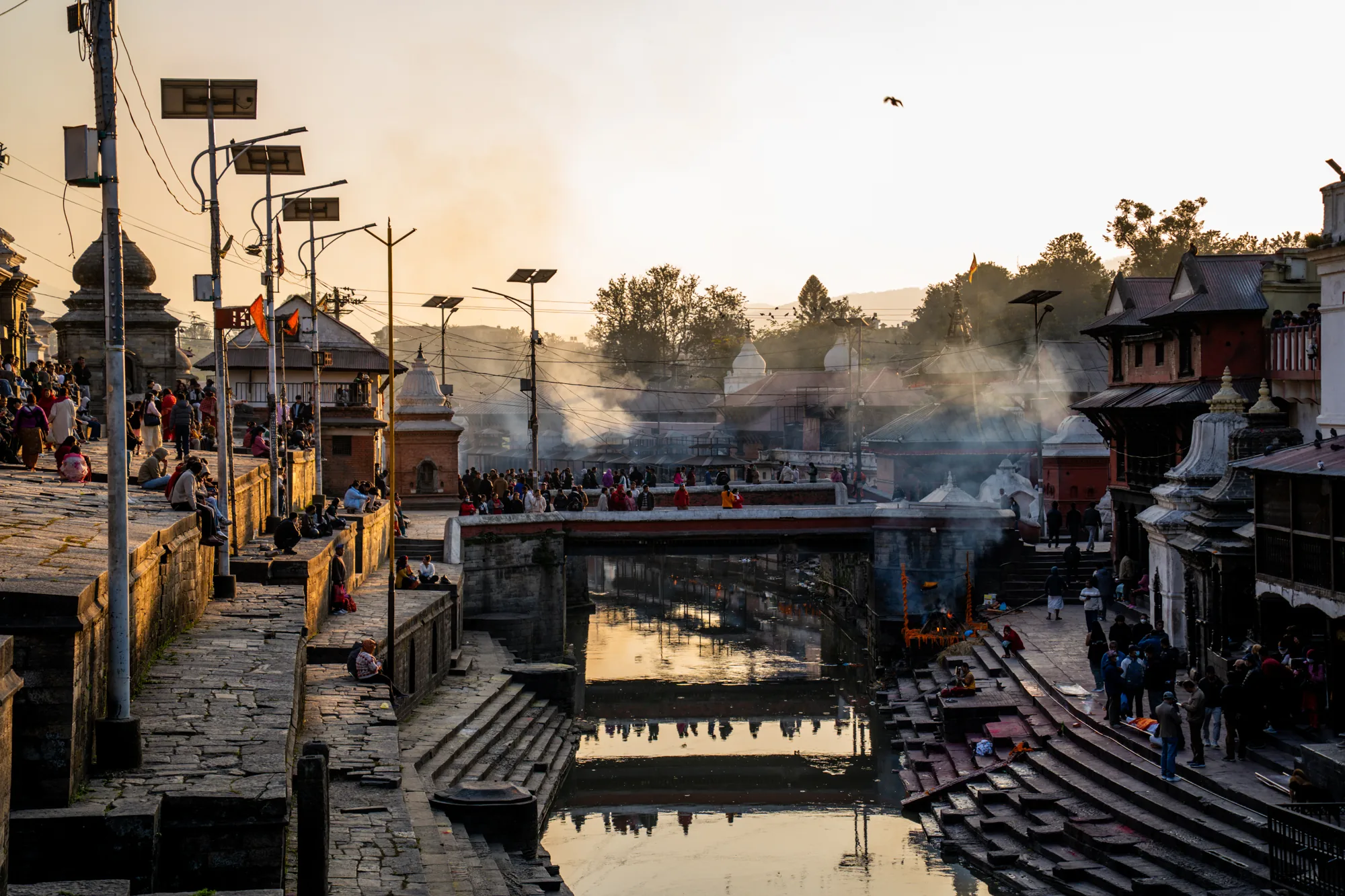 この写真はネパールのパシュパティナートで撮影されたもので、黄金色の夕暮れ時、聖なるバグマティ川とその周辺の風景が写されています。川の両岸には石造りの階段や建造物が立ち並び、多くの人々が川沿いに座ったり、歩いたりしています。川には橋が架かっており、橋の上にも人が集まっているのが見えます。  背景には、煙が立ち上っているのが見えます。これは、この場所が火葬場としても使われているためでしょう。写真全体は、穏やかで神聖な雰囲気に包まれており、人々の日常生活と宗教的な儀式が共存する様子が捉えられています。川面に映る建物の影や、夕日に照らされた石造りの階段が、独特の美しさを添えています。空には鳥が飛んでおり、全体的に静かで平和な印象を与える写真です。