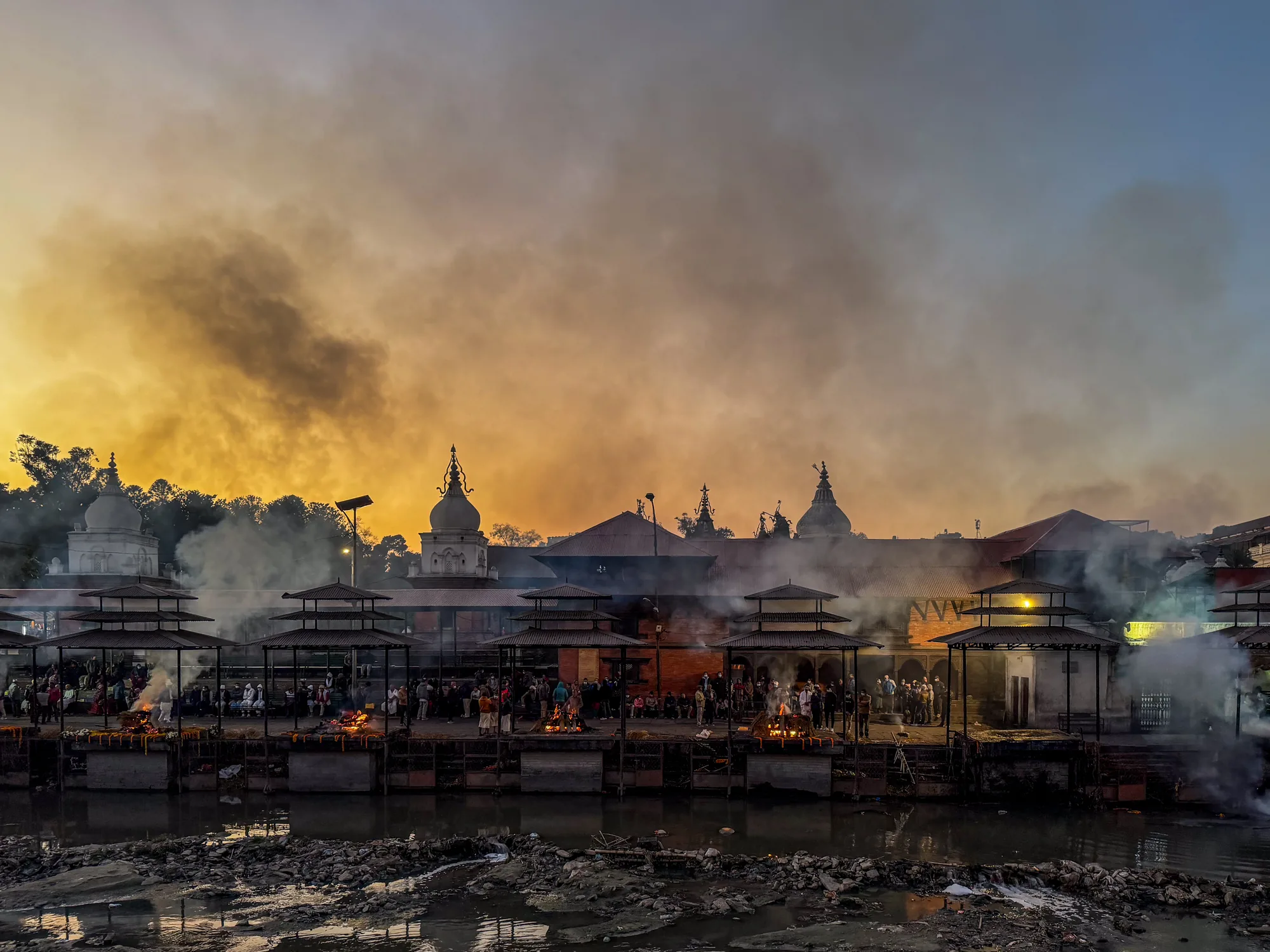 この写真はネパールのパシュパティナートで撮影されたものです。手前には汚れた川が流れ、その川沿いに火葬場が並んでいます。火葬場では火が燃え盛っており、立ち上る煙が空を覆っています。多くの人々が集まっており、それぞれの場所で故人を弔っているようです。背景には寺院の建物がそびえ立ち、夕焼け空が煙に染まり、独特の雰囲気を醸し出しています。全体的に、死と生が交錯する、厳かで神秘的な光景が捉えられています。