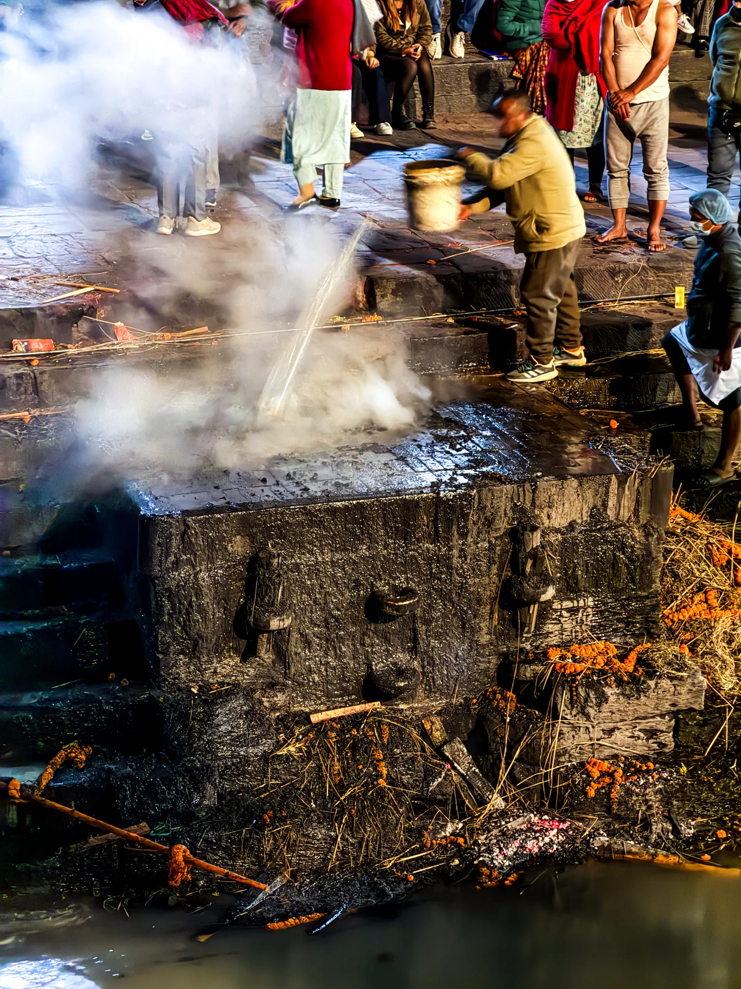 ネパールのパシュパティナートで撮影された写真です。写真には、火葬の儀式が行われている様子が写っています。中央には、燃え尽きた薪の残骸が積み上げられた石造りの祭壇が見え、その上から男性がバケツで水をかけています。水蒸気が激しく立ち上り、あたりを白く霞ませています。祭壇の下には川が流れ、川岸には燃え残った薪やマリーゴールドの花飾りが散乱しています。祭壇の周りには、儀式の様子を見守る人々が立っています。彼らの服装は様々で、日常着を着た人もいれば、伝統的な衣装を身につけた人もいます。写真全体は、厳かで神秘的な雰囲気を醸し出しています。光の具合と立ち上る水蒸気により、現実と非現実が混ざり合ったような独特な空気感が表現されています。
