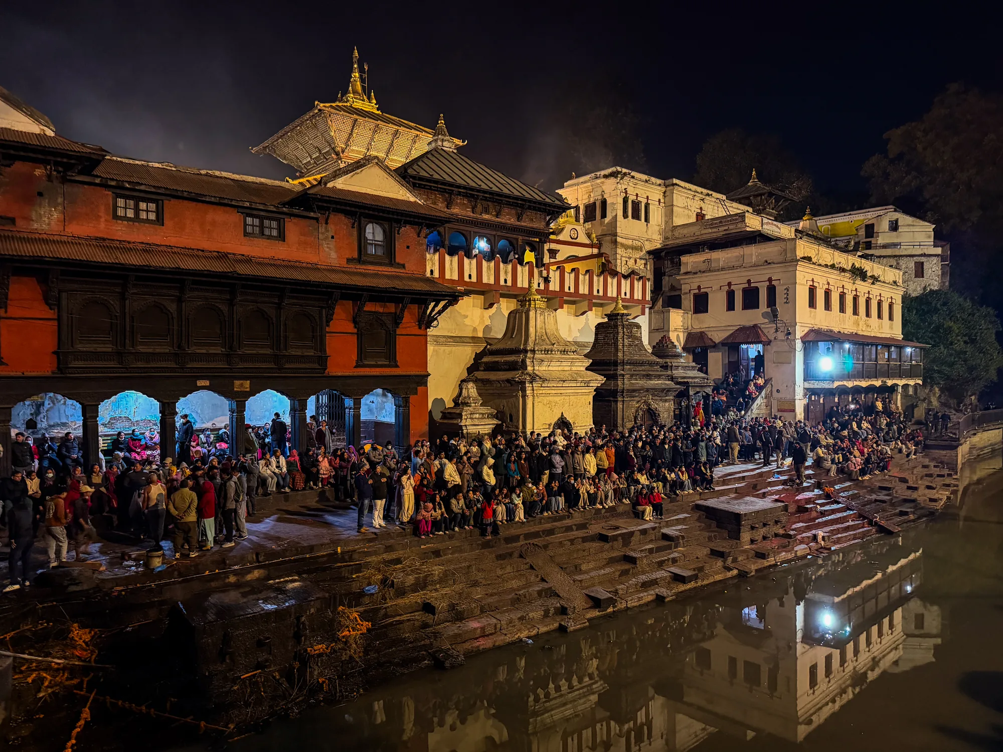 写真はネパールのパシュパティナートで夜間に撮影されたものです。画面中央にはバグマティ川が流れ、その両岸には多くの人々が集まっています。川岸には階段状の石組みがあり、人々はそこに座ったり立ったりして、何かの儀式やイベントを見ているようです。川の水面には、建築物の灯りが反射して揺らめき、幻想的な雰囲気を醸し出しています。  川の背景には、歴史を感じさせる建築物が立ち並んでいます。赤茶色や白を基調とした建物は、伝統的なネパールの様式で、細かな装飾が施されています。特に目を引くのは、中央奥にある金色の屋根を持つ寺院で、その荘厳な姿は神聖な場所であることを示しています。  写真全体は夜の暗さを基調としていますが、建築物や人々の灯りによって照らされ、温かい光が差し込んでいます。多くの人々が集まる様子や、建築物の美しさ、そして川面に映る光景が一体となり、パシュパティナートの神秘的な雰囲気を伝えています。夜の静けさの中で、人々の祈りや信仰が感じられるような、印象的な一枚です。