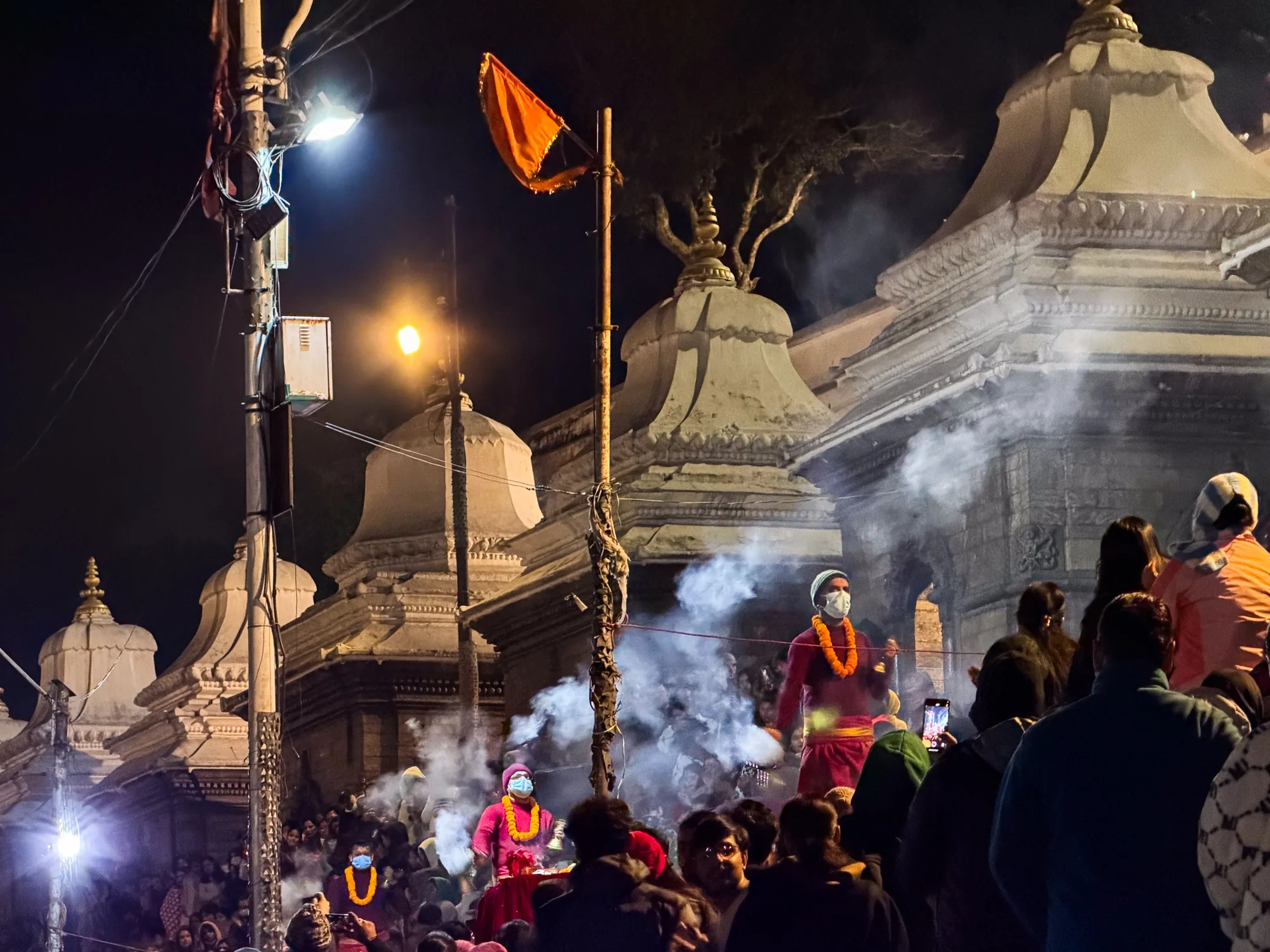 ネパールのパシュパティナートで撮影された夜の風景です。暗闇の中にライトが灯り、寺院の白い建物が浮かび上がっています。煙が立ち込め、幻想的な雰囲気を醸し出しています。  大勢の人々が集まり、祭りのような賑わいを見せています。オレンジ色の衣装を着た人物が複数見られ、首にはマリーゴールドのネックレスをかけています。マスクを着用している人もおり、コロナ禍の状況がうかがえます。  旗がはためき、人々はスマートフォンで写真を撮っています。神聖な場所で、人々の信仰心や熱気が伝わってくるような一枚です。