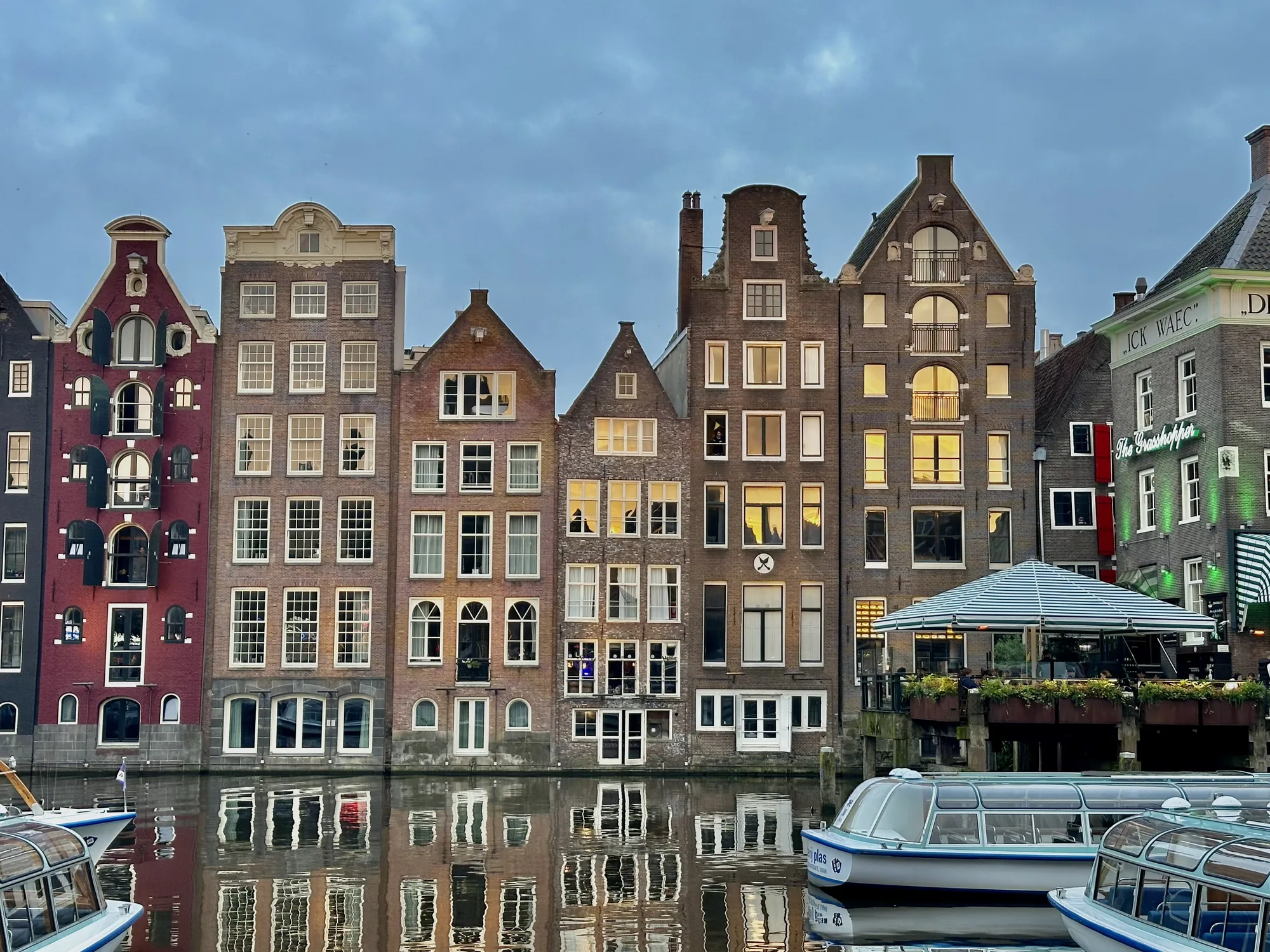 写真はオランダのアムステルダムで撮影された、運河沿いの風景を捉えたものです。歴史を感じさせる建物が運河に沿って立ち並び、水面に映る建物や窓の光が幻想的な雰囲気を醸し出しています。空は曇り空で、夕暮れ時か夜明け前の薄明かりの中、建物内部の明かりが暖かく灯っています。  特に目を引くのは、個性的なデザインの建物群です。レンガ造りの建物はそれぞれ異なる形状の屋根を持ち、窓の配置や装飾も様々で、独特の景観を作り出しています。一番左の建物は赤色で目立ち、その隣には多くの窓が並んだ建物が続いています。いくつか窓が開いていたり、人が見える窓もあります。  運河には観光用のボートが浮かんでおり、静かな水面をゆっくりと進んでいる様子が想像できます。ボートの色合いと運河沿いの建物が調和し、絵画のような美しさを生み出しています。運河沿いにはテラス席のあるレストランも見え、人々が食事を楽しんでいる様子もうかがえます。全体的に落ち着いた色調でまとめられており、アムステルダムの穏やかな雰囲気が伝わってくる写真です。