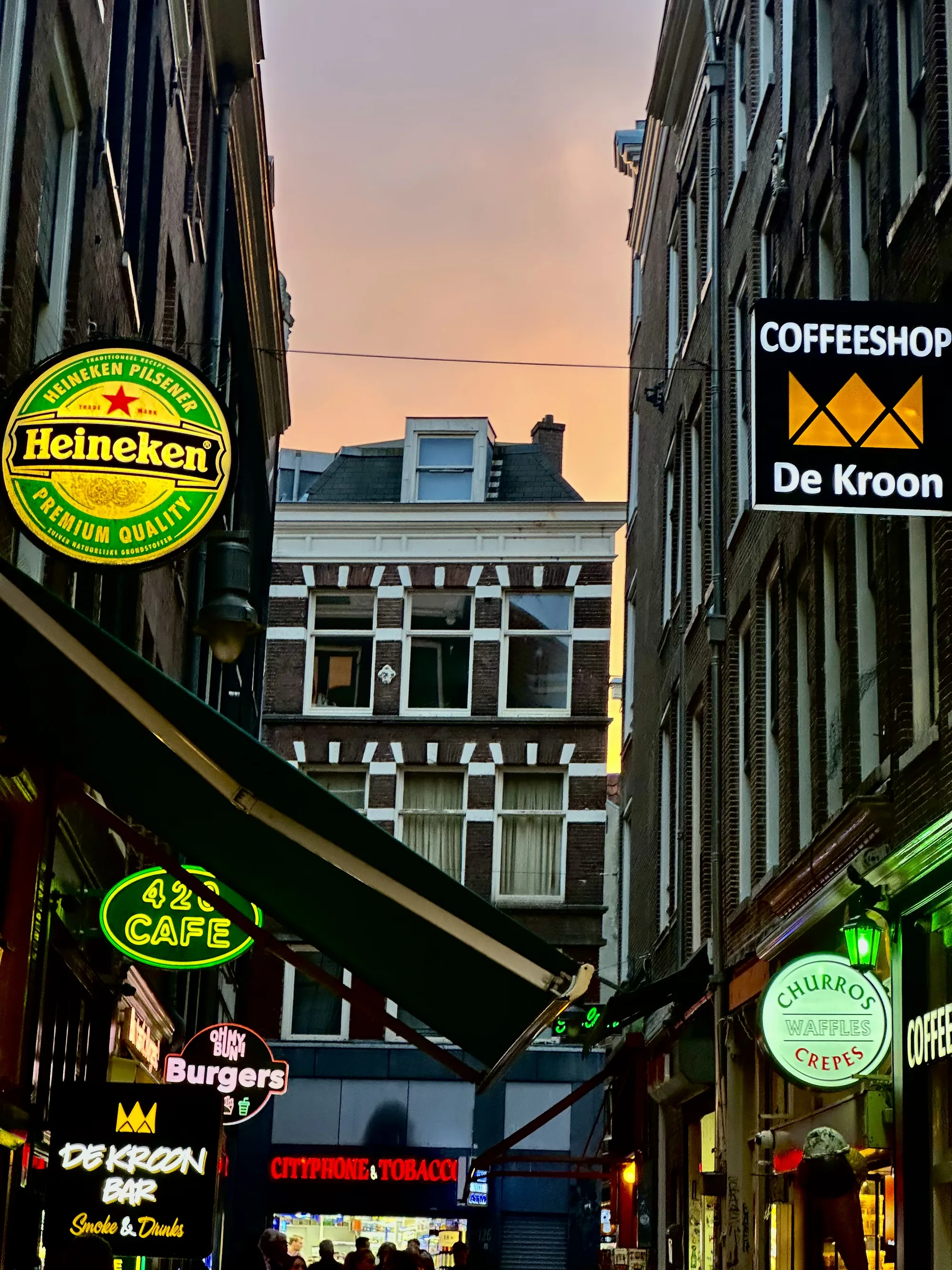 この写真は、オランダのアムステルダムの路地裏を捉えたものです。両側にレンガ造りの建物が立ち並び、狭い道の上には電線が走っています。空は夕焼けの色に染まり、建物と建物の間から見える夕暮れの光が美しいコントラストを生み出しています。  道の両側には、様々な店舗の看板が掲げられています。ハイネケンの大きな看板、420 CAFEのネオンサイン、Burgersという文字、COFFEESHOPの看板、そして「De Kroon Bar Smoke & Drinks」と書かれた看板など、多種多様な店が存在することがわかります。また、「CHURROS Waffles Crepes」と書かれた看板もあり、軽食を提供する店もあるようです。これらの看板は、活気のある雰囲気を醸し出しています。  画面下部には、複数の人の姿が見えます。彼らは店の前を歩いていたり、店の中に入ろうとしていたりするようです。写真全体から、アムステルダムの賑やかな夜の街の様子が伝わってきます。観光客だけでなく、地元の人々も集まる、親しみやすい雰囲気の場所であることがうかがえます。