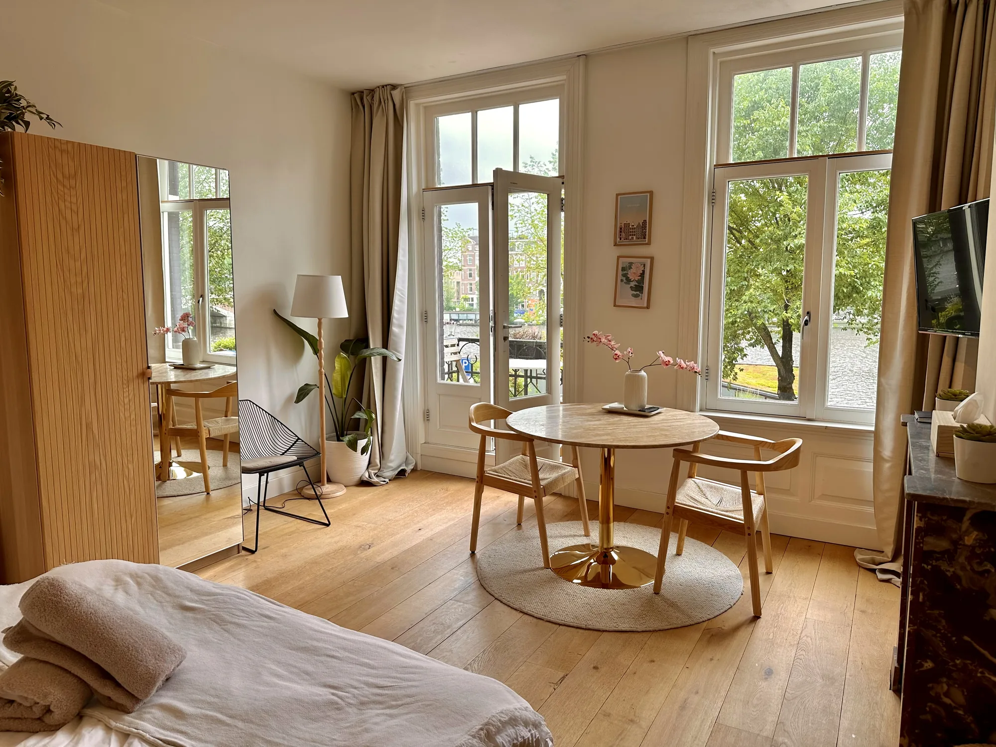 オランダ、アムステルダムで撮影された室内の写真です。部屋全体は、明るく温かみのある雰囲気で、木製のフローリングが特徴的です。写真手前にはベッドの一部が写っており、ベージュ色のブランケットが畳まれています。部屋の中央には、丸いテーブルと2脚の椅子が配置され、テーブルの上には花瓶に入ったピンクの花が飾られています。窓からは運河が見え、アムステルダムらしい景観が楽しめます。部屋には、観葉植物やスタイリッシュな椅子、木製のキャビネットなどが配置され、洗練されたインテリアが印象的です。全体的に、落ち着いた雰囲気で、快適な空間が演出されています。
